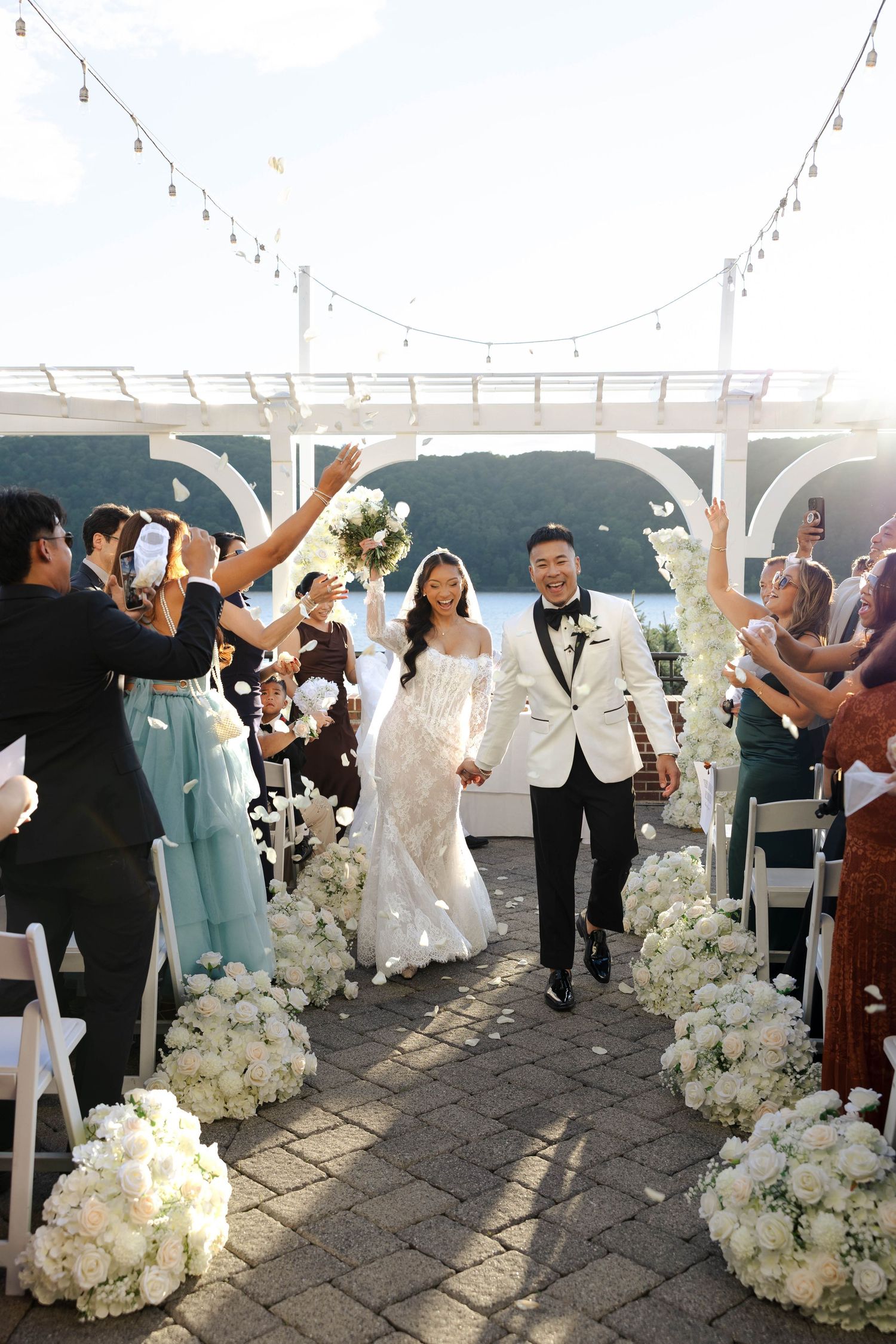 Bride and groom walk down outdoor aisle lined with white floral arrangements, guests celebrating, waterfront venue backdrop.