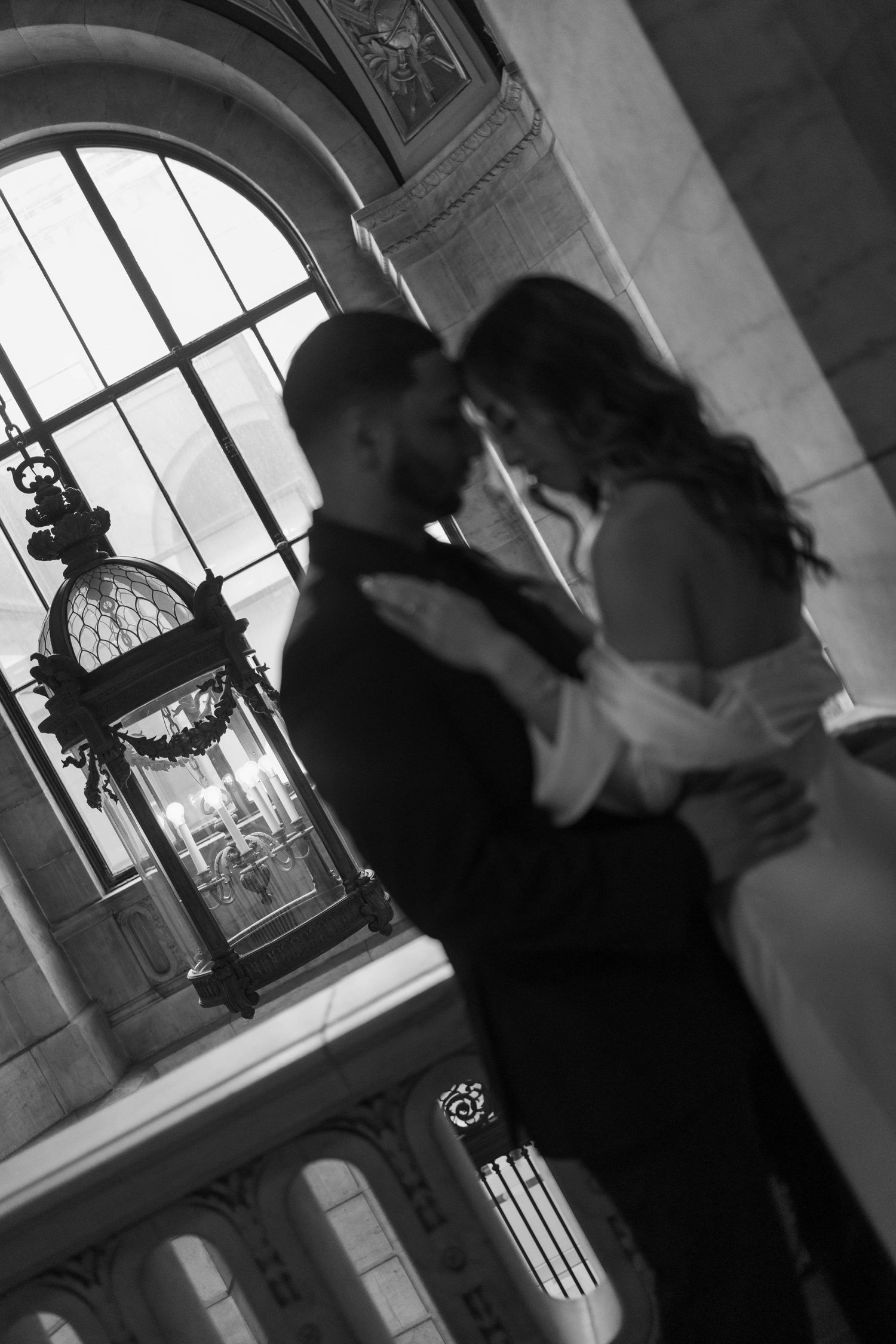 Black and white romantic portrait of bride and groom holding each other near ornate arched window.