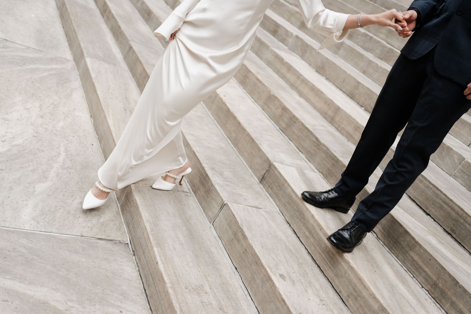 Close-up of bride in white heels and groom in black dress shoes on marble steps.