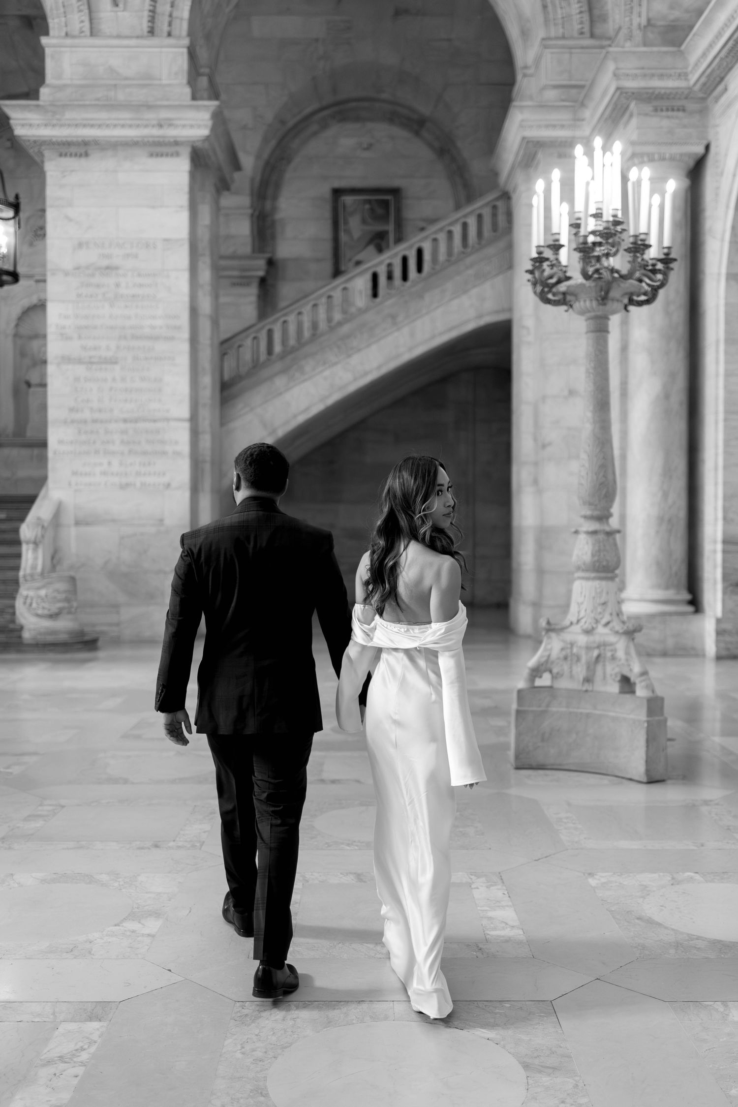 Black and white photo of bride and groom walking past a candelabra in a grand marble interior.