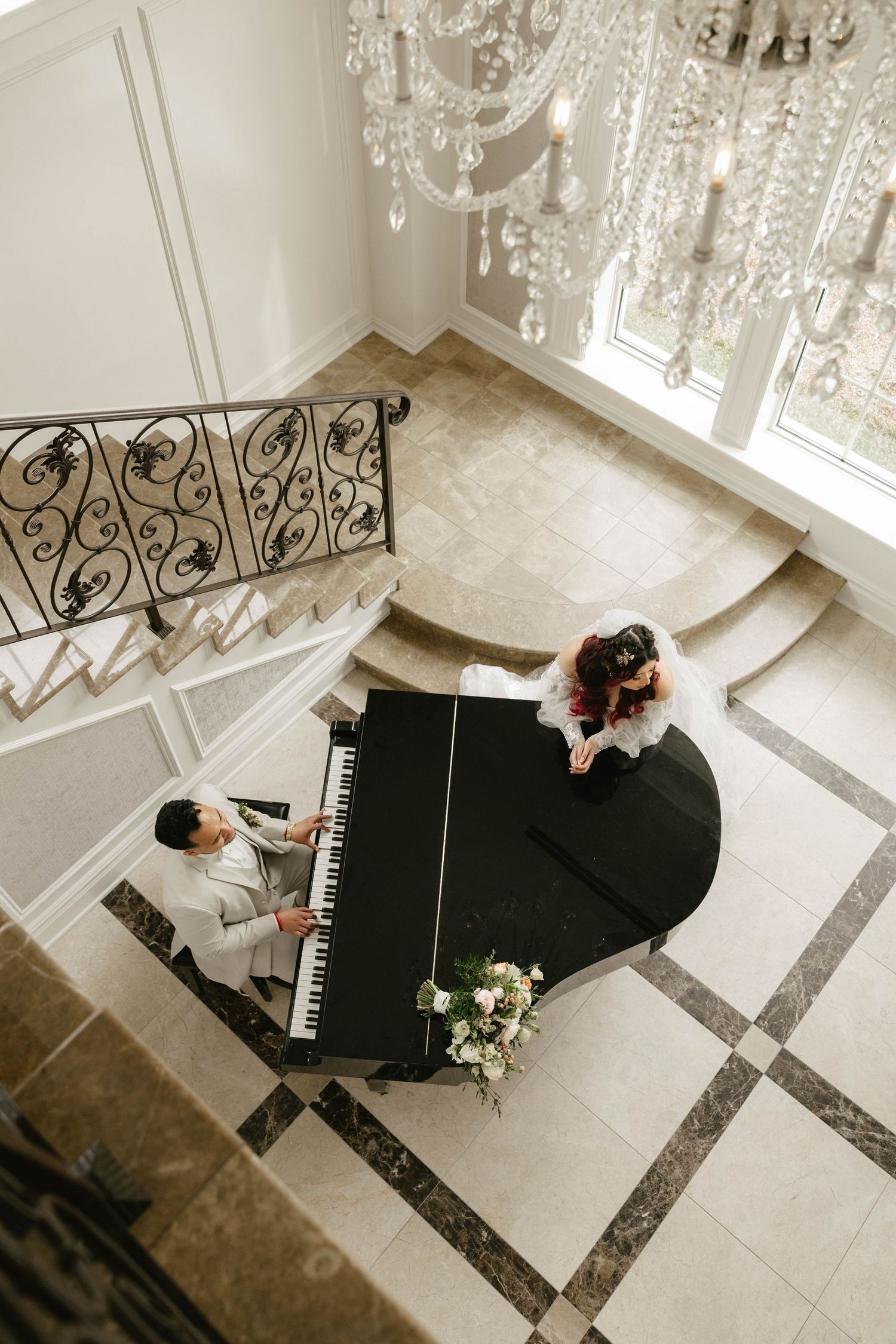 The Aria Wedding & Banquet Facility - Overhead view of groom playing grand piano for bride in elegant marble foyer with crystal chandelier.