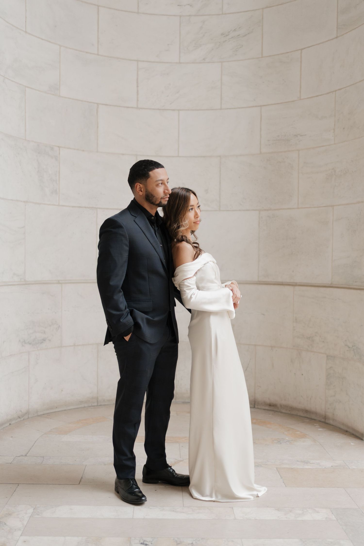 Couple in wedding attire sharing intimate moment against curved marble wall interior.