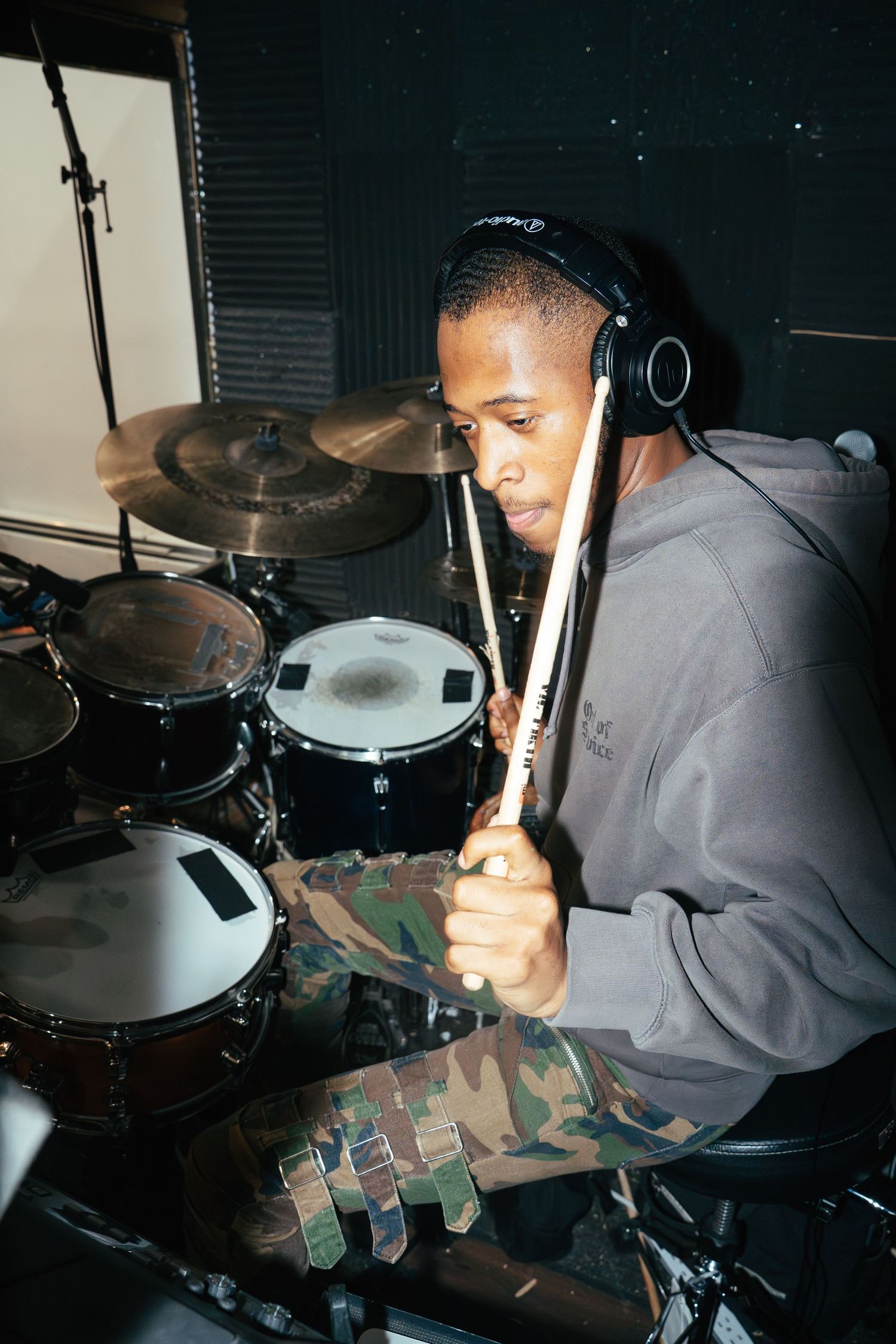 A drummer performs in a dark studio setting wearing headphones and a camouflage pattern shirt.