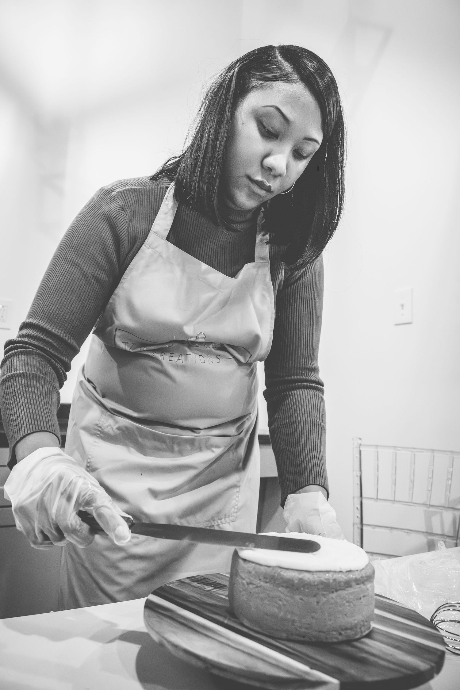 A person in an apron slices a round cake with a large knife on a white surface.