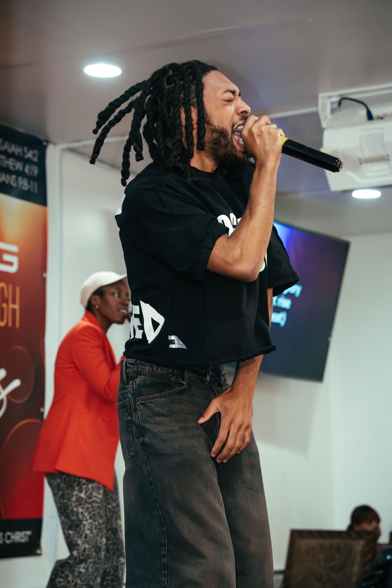 A performer with dreadlocks sings into a microphone wearing a black t-shirt on stage.