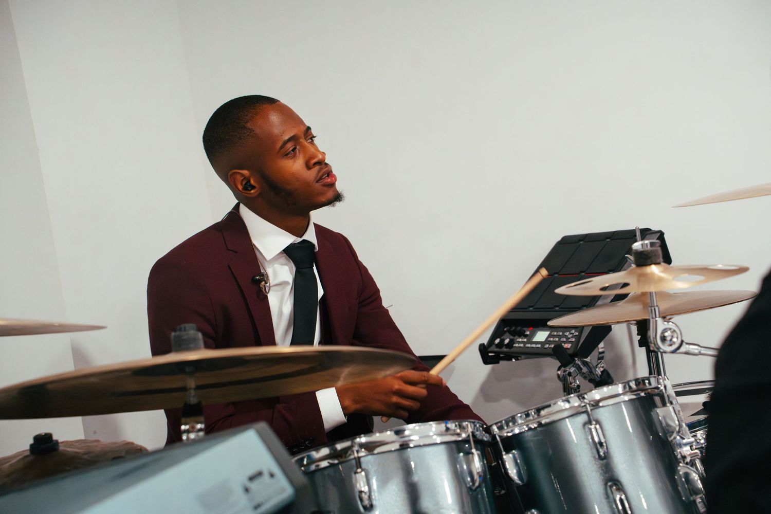 Professional musician plays a silver drum set while wearing a burgundy suit jacket and black tie.