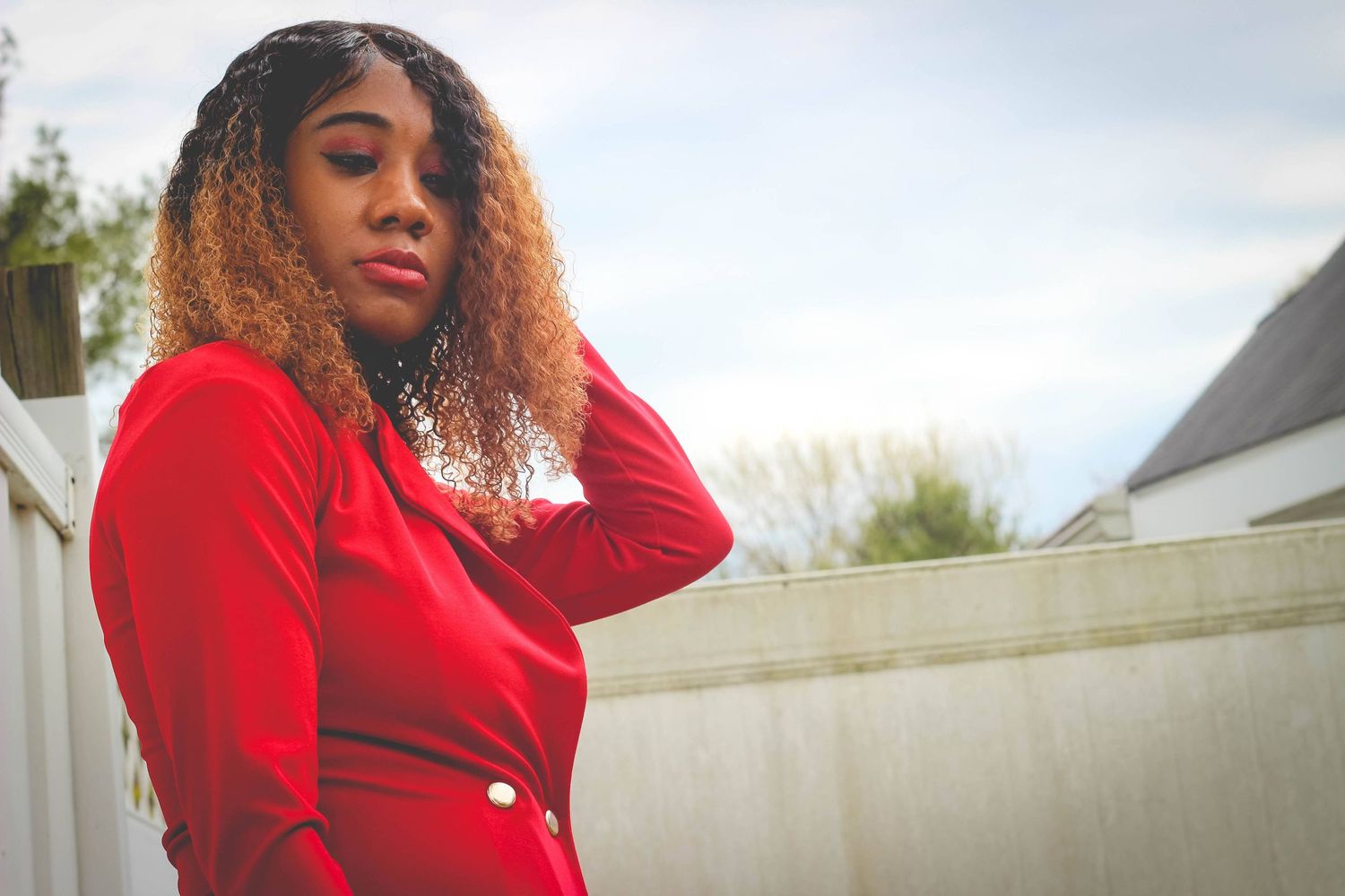 Person with curly hair wearing a vibrant red sweater poses outdoors against a white wall in natural lighting.