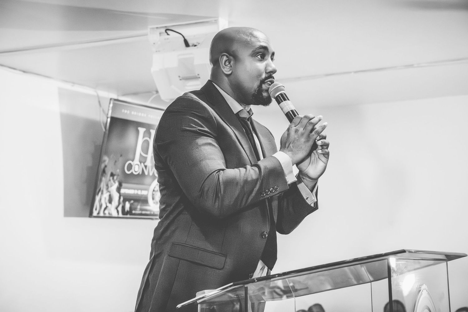 A speaker in a dark suit gestures while presenting at a business event or conference.