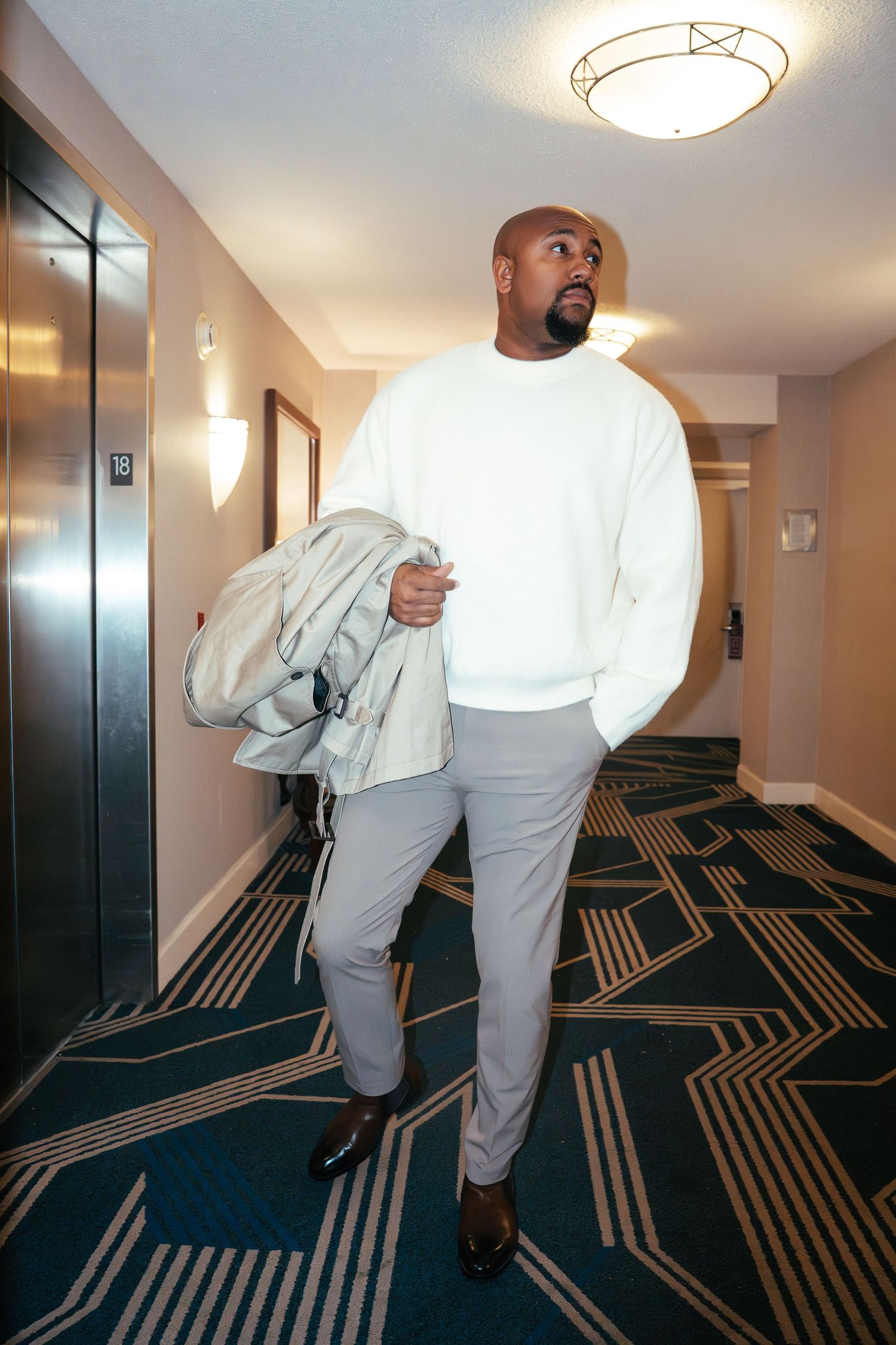 A person in a white sweater and gray pants stands in a hotel hallway with geometric carpet.