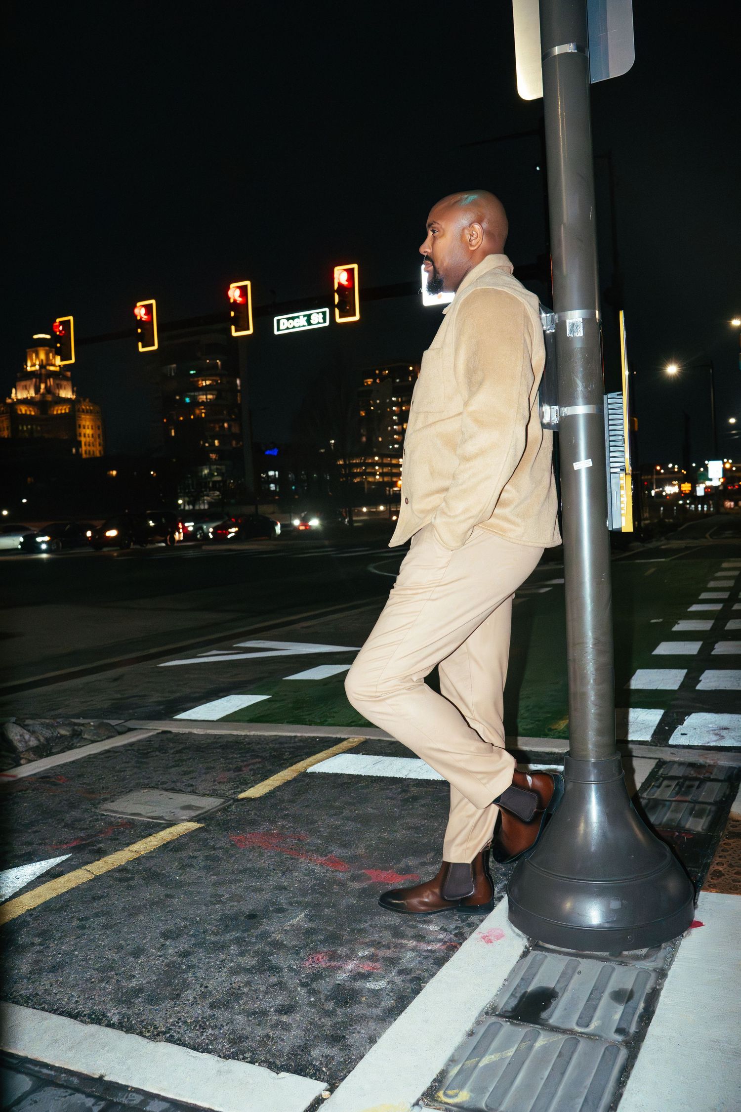 Person in beige suit standing on city street corner at night with illuminated buildings in background.
