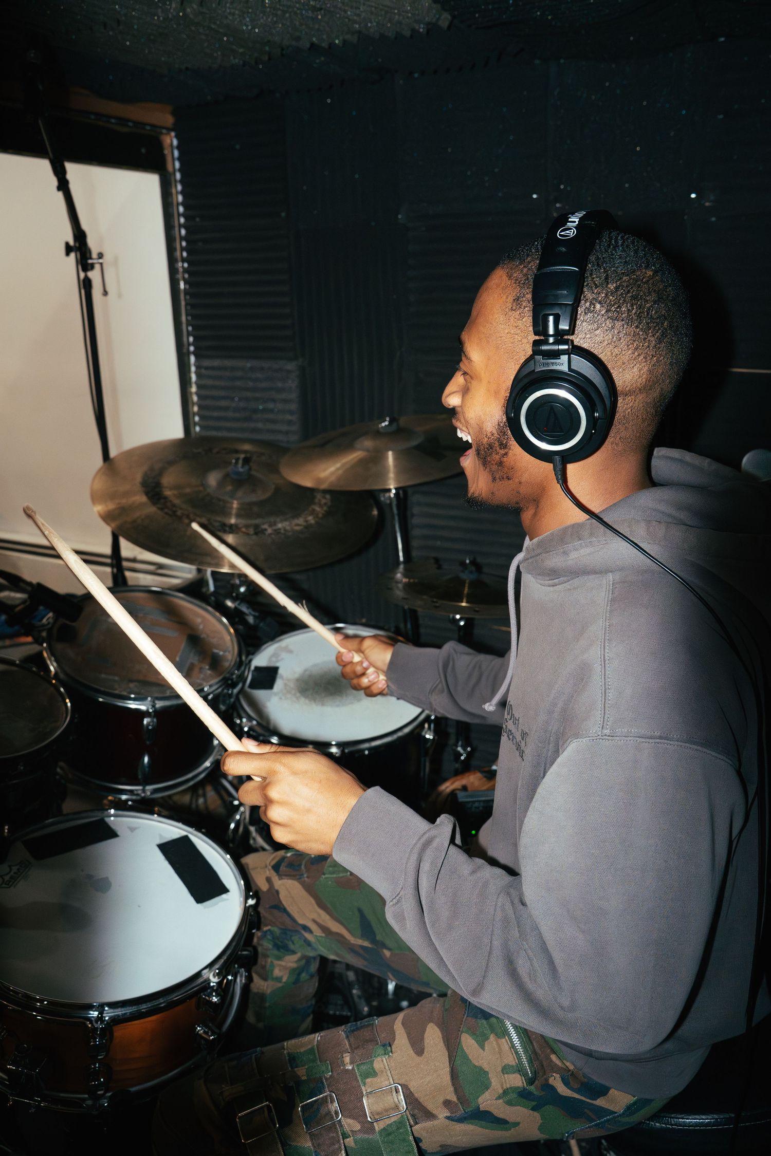 A drummer in a grey hoodie plays an electronic drum kit while wearing professional headphones.