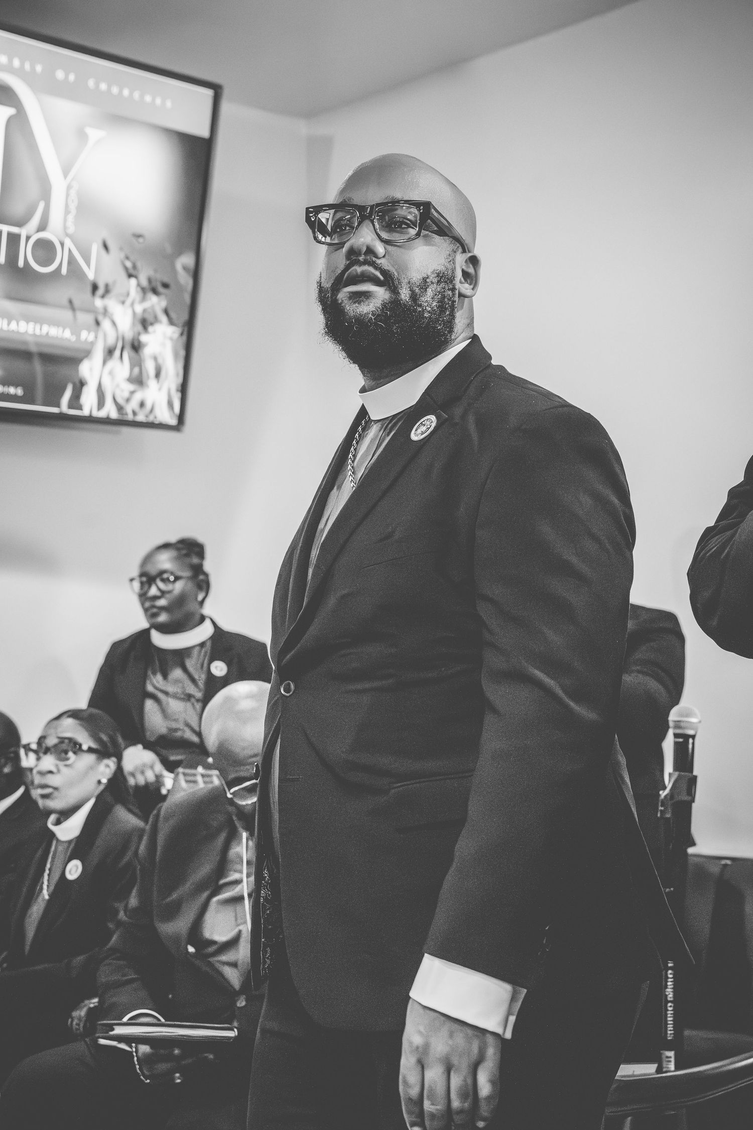 A speaker in a black suit and glasses stands during a formal event.