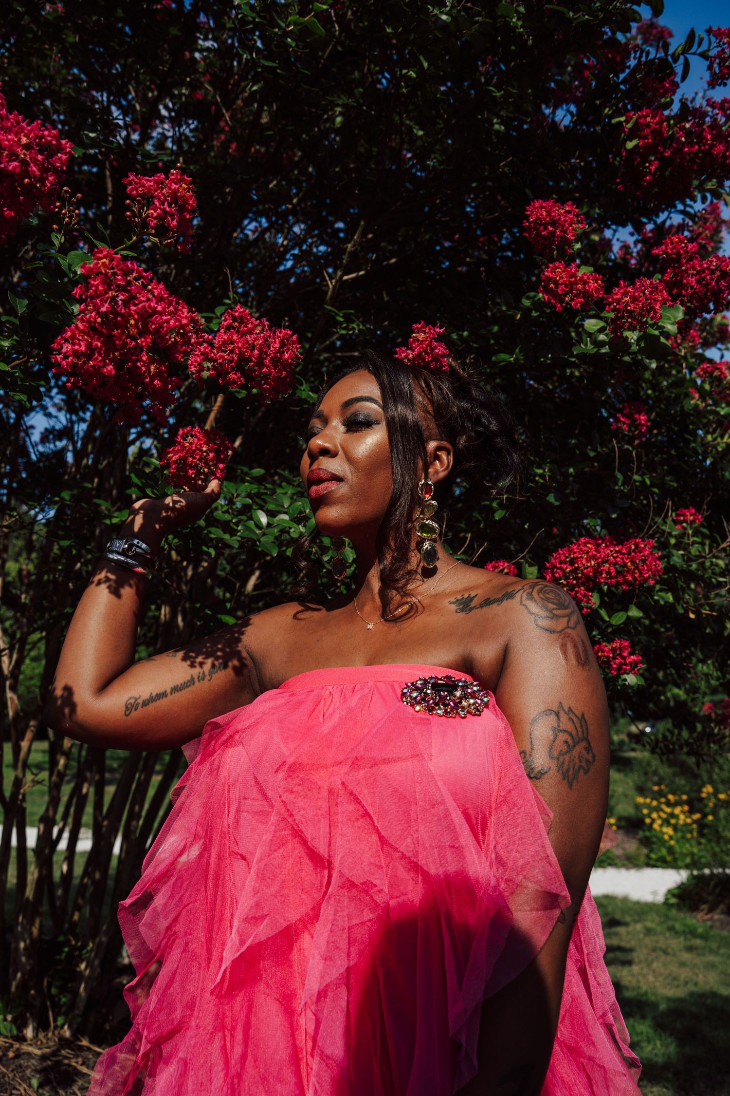 A stunning portrait in front of blooming red flowers features a pink tulle gown and artistic tattoos in golden sunlight.