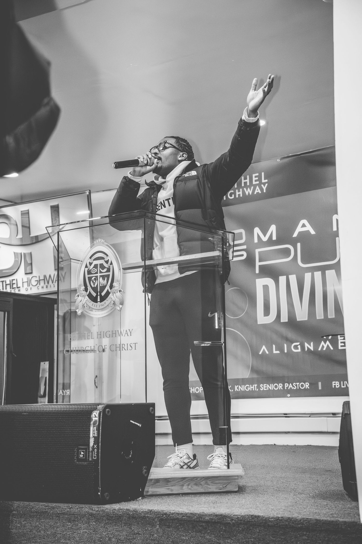 Black and white photo of performers with raised arms on stage with branded banners in background.