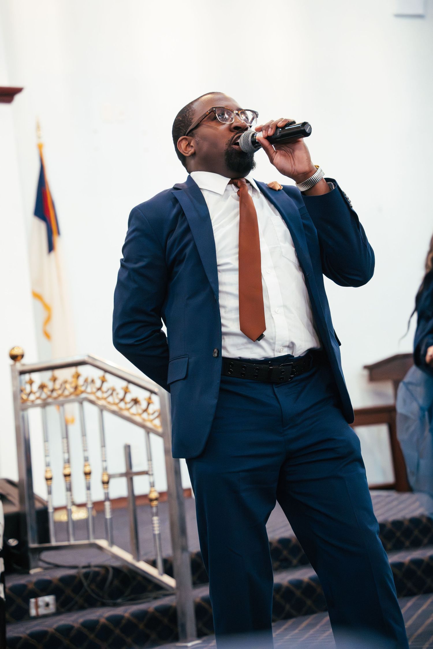 A performer in a navy suit and brown tie sings into a microphone while standing on a stage.