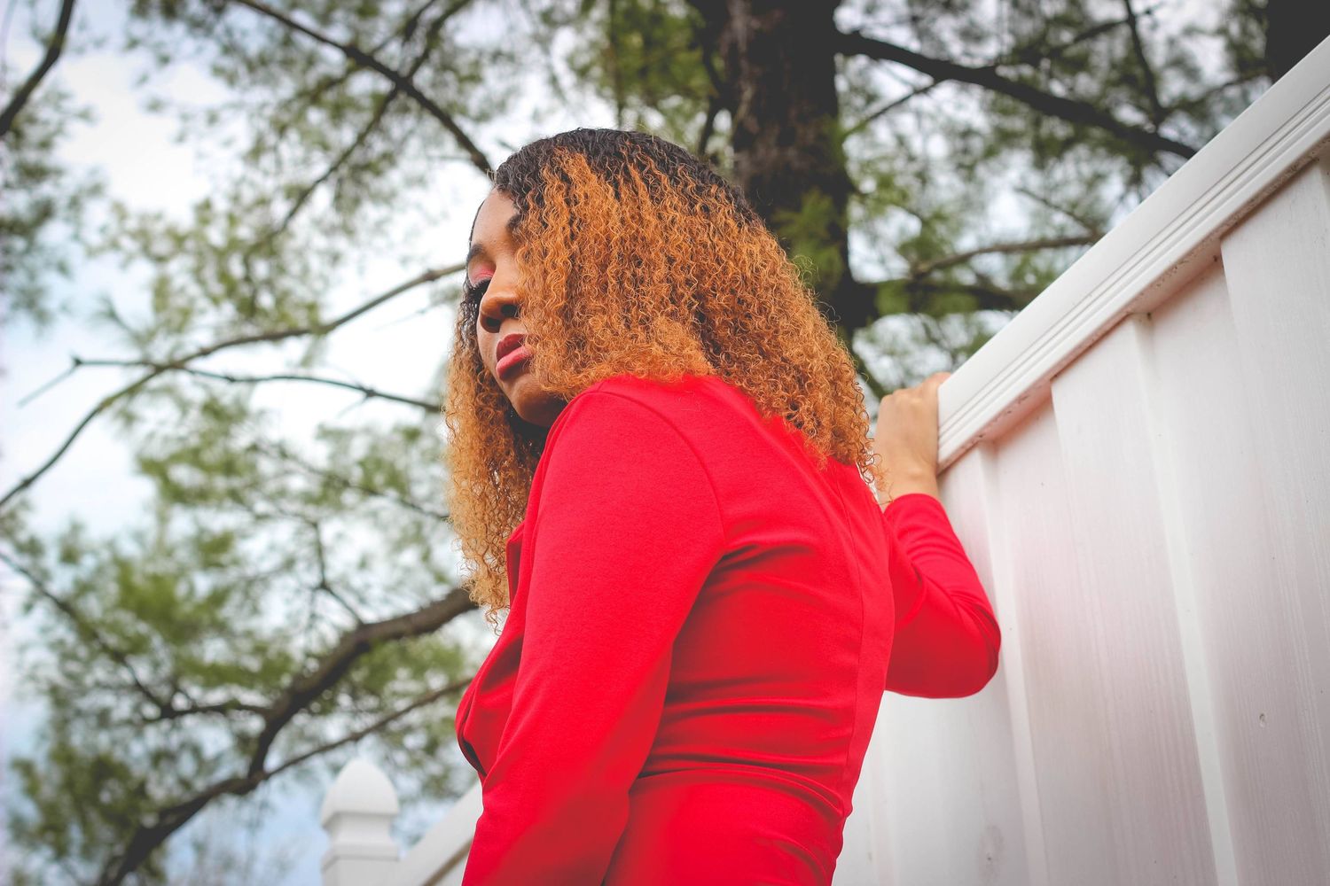 A person in a bright red sweater stands against a white wall with trees visible in the background.