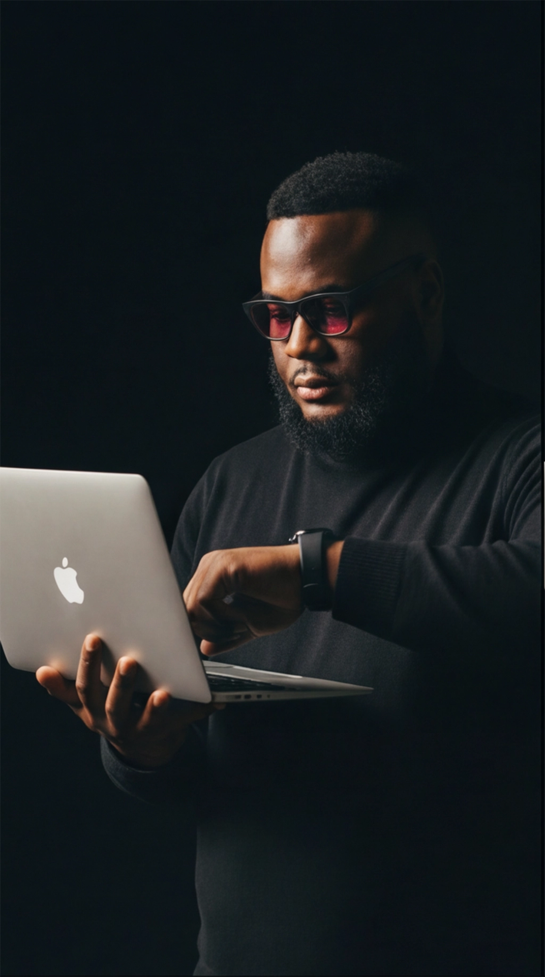 Person in dark room working on MacBook laptop with dramatic lighting creating moody atmosphere.