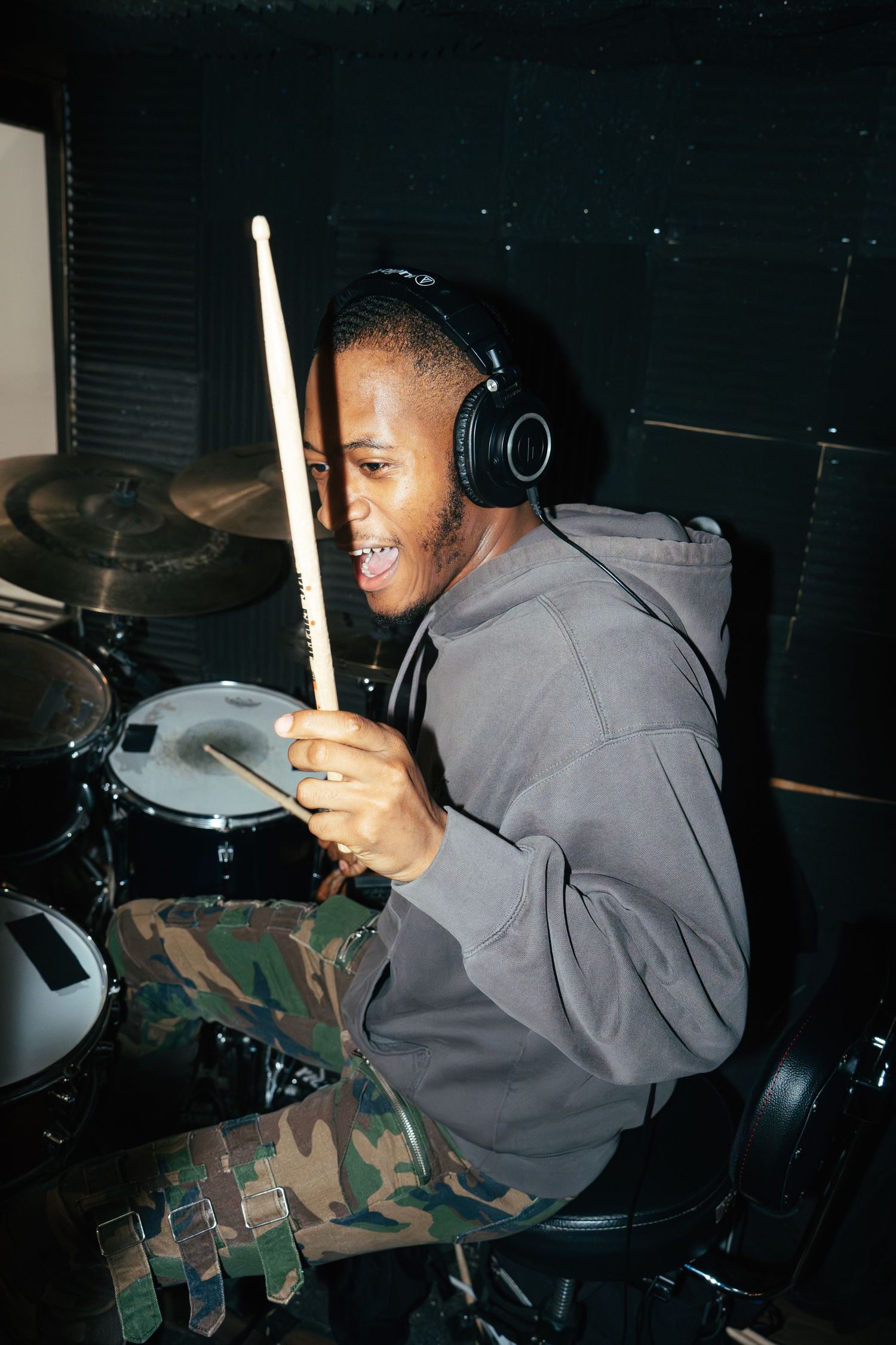 A drummer wearing headphones plays drums with a smile in a dark studio setting.