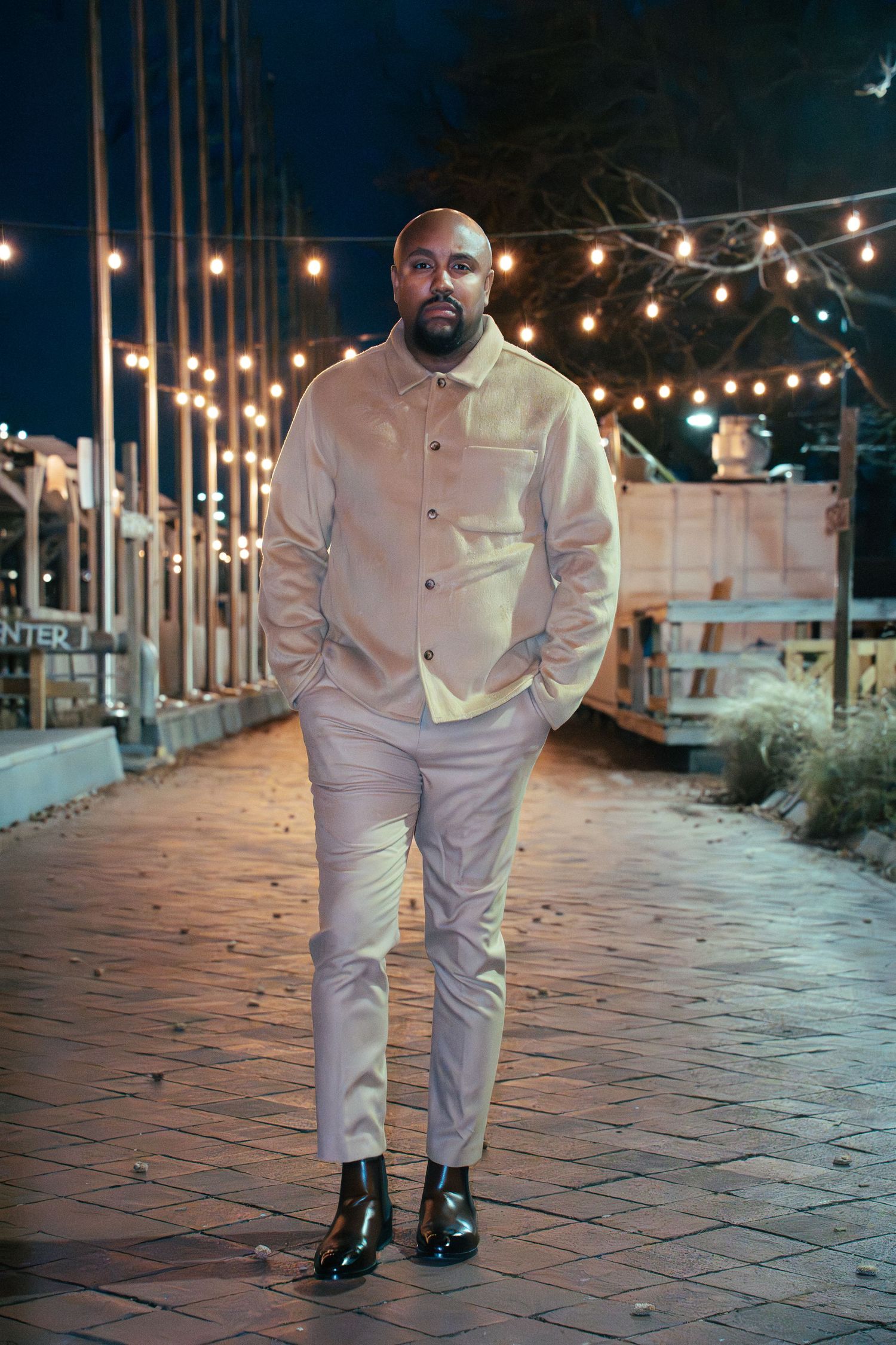 A person in a beige outfit stands on a brick walkway at night with string lights glowing overhead.