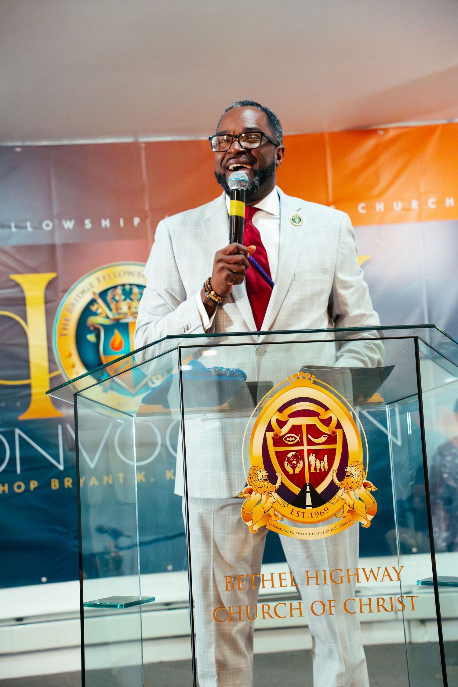 A speaker in a light suit and red tie addresses an audience from behind a glass podium with a church logo.