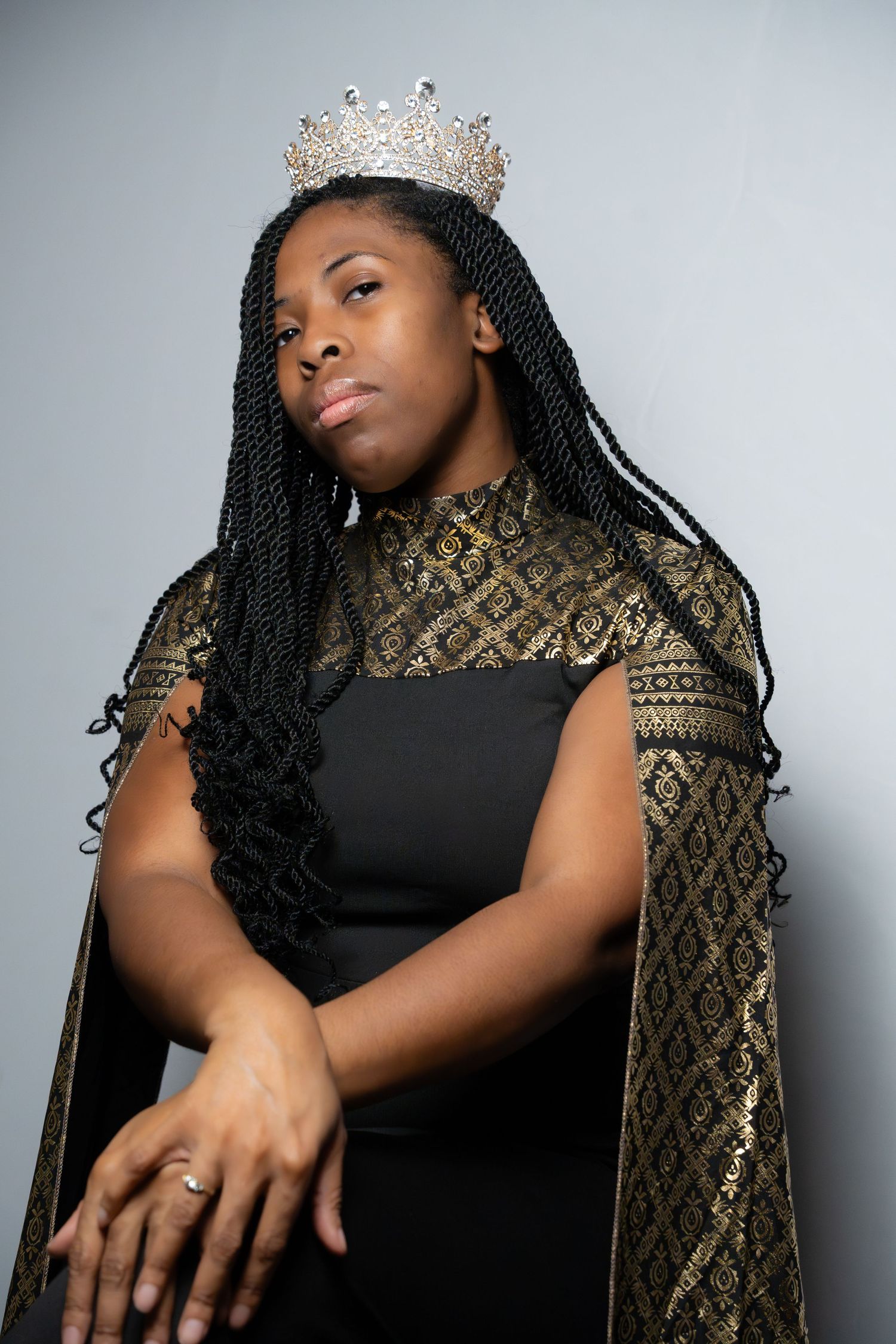 A regal portrait featuring a sparkling crown and black and gold formal attire against a gray backdrop.