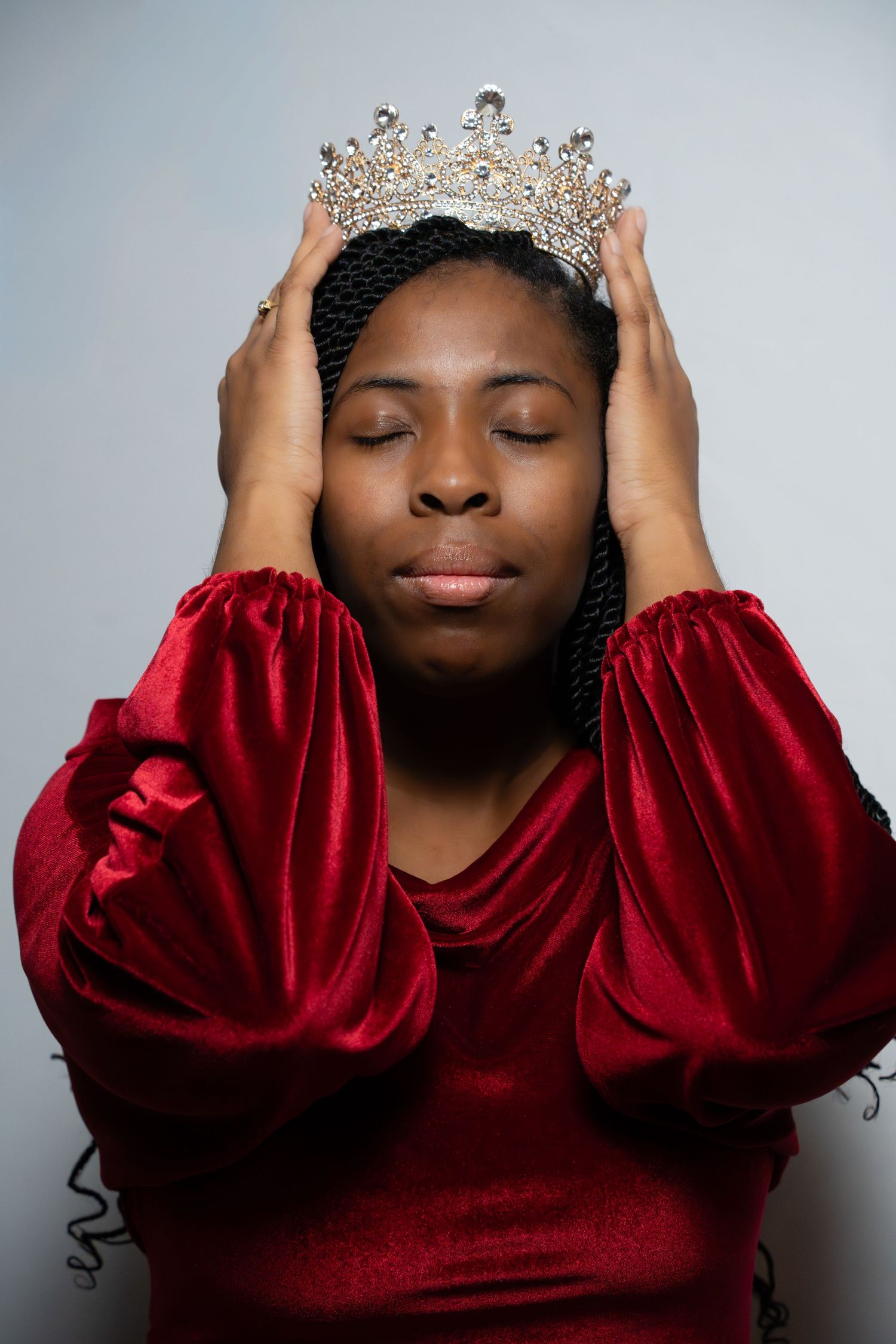 A person wearing a red velvet dress places a sparkling crown on their head against a gray background.