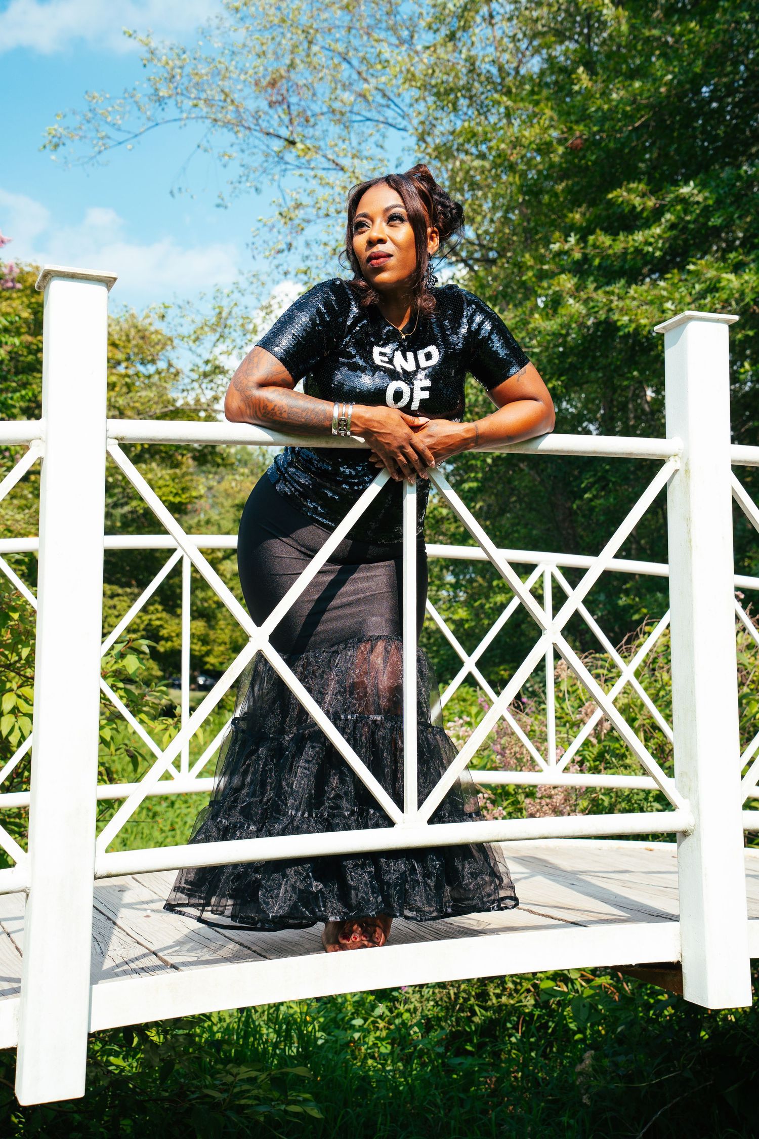A figure in a black t-shirt and long skirt poses on a white railing outdoors with trees in the background.