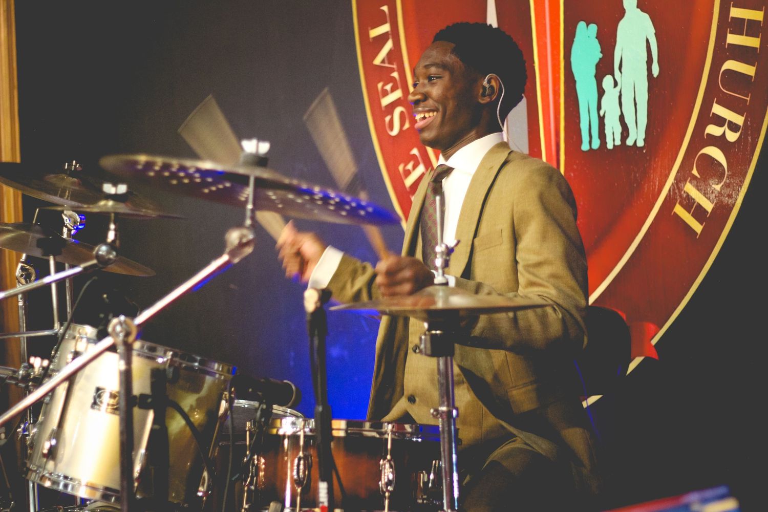 Drummer in tan suit performing energetically behind drum kit at jazz club with red wall and neon decor.