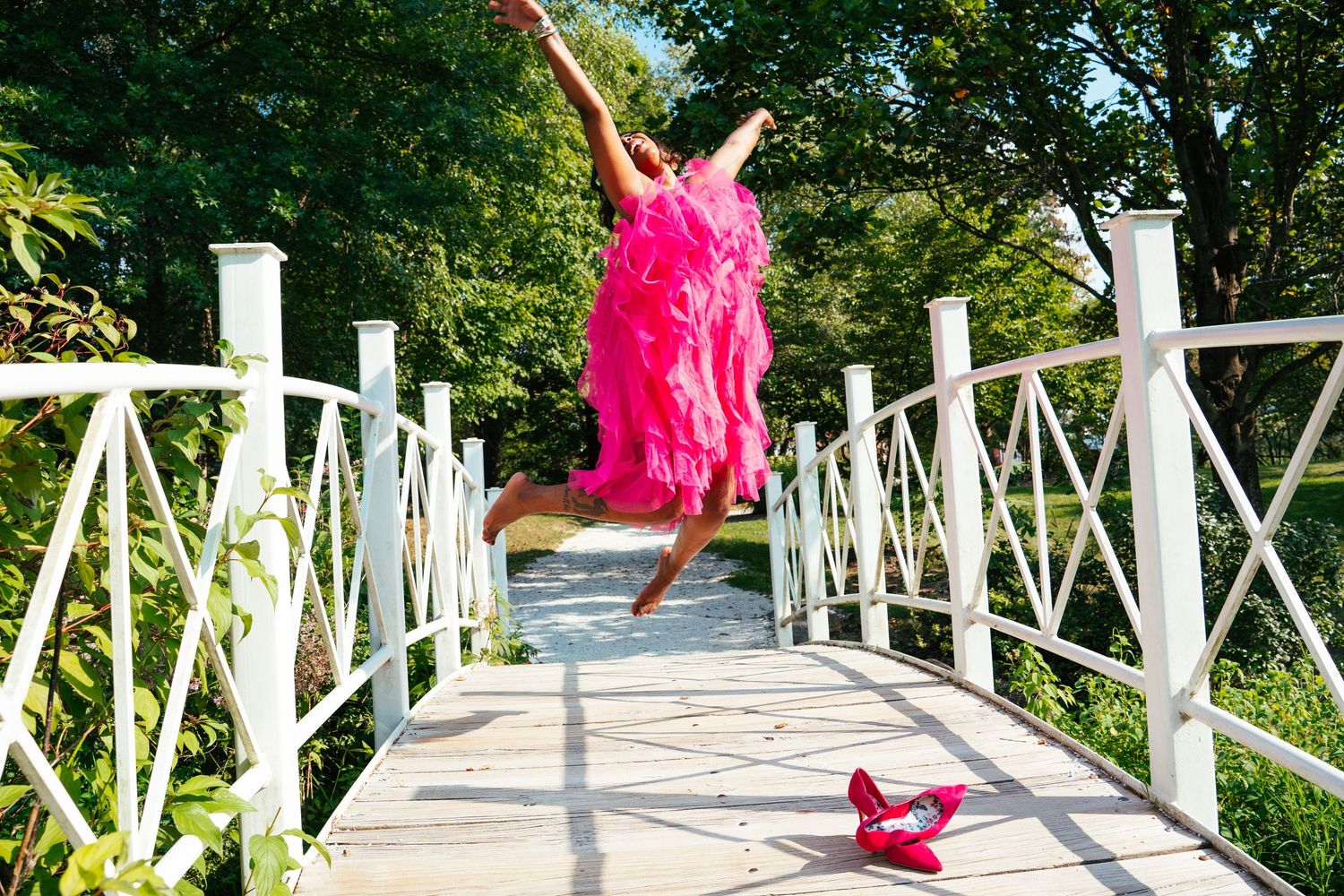 Figure in bright pink dress jumps joyfully on a white wooden bridge with red shoes visible below.