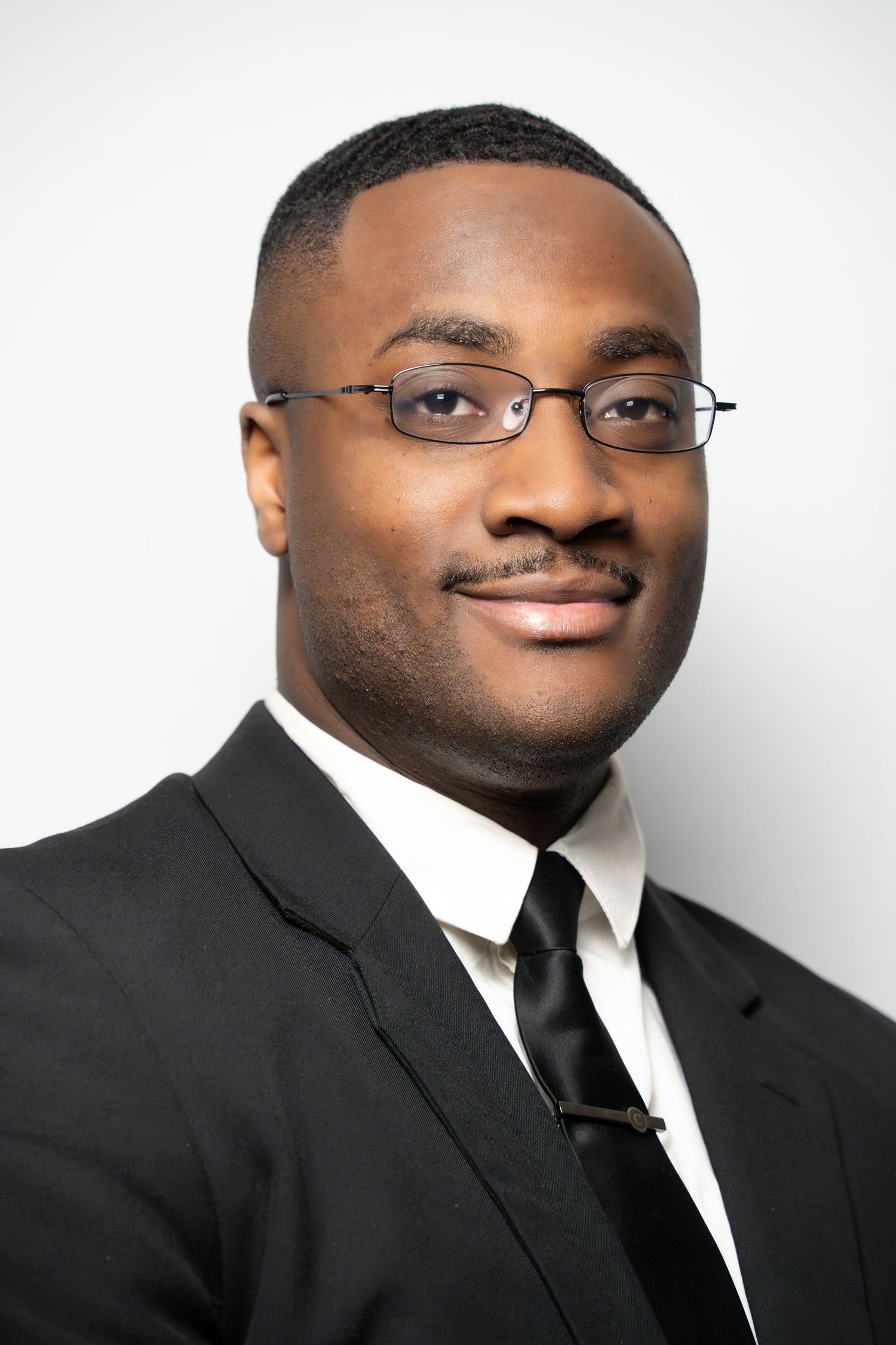 Professional headshots of a person wearing glasses and a black suit with white shirt and tie against a light background.