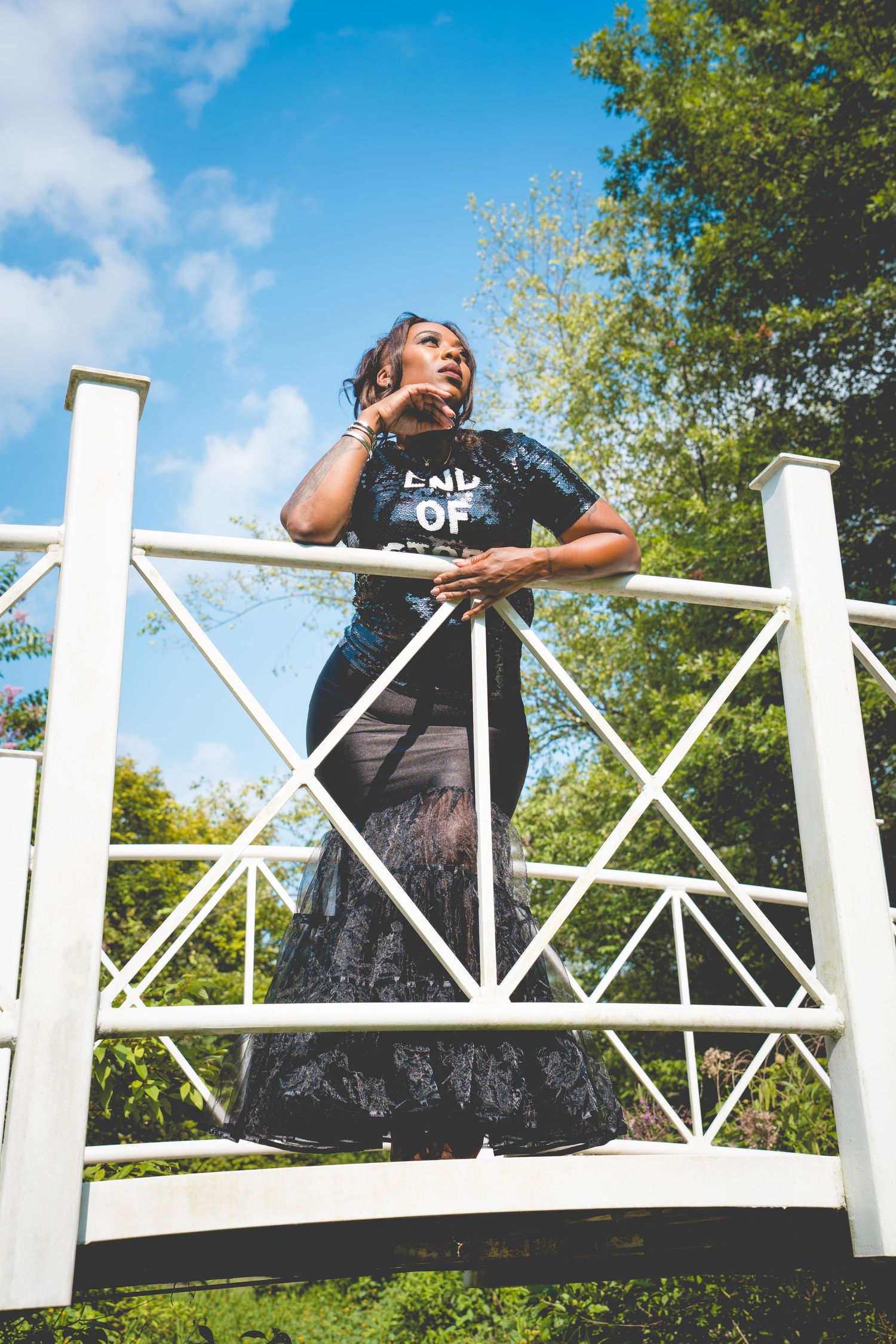 A figure stands on a white decorative bridge railing wearing a black t-shirt against a blue summer sky with trees.
