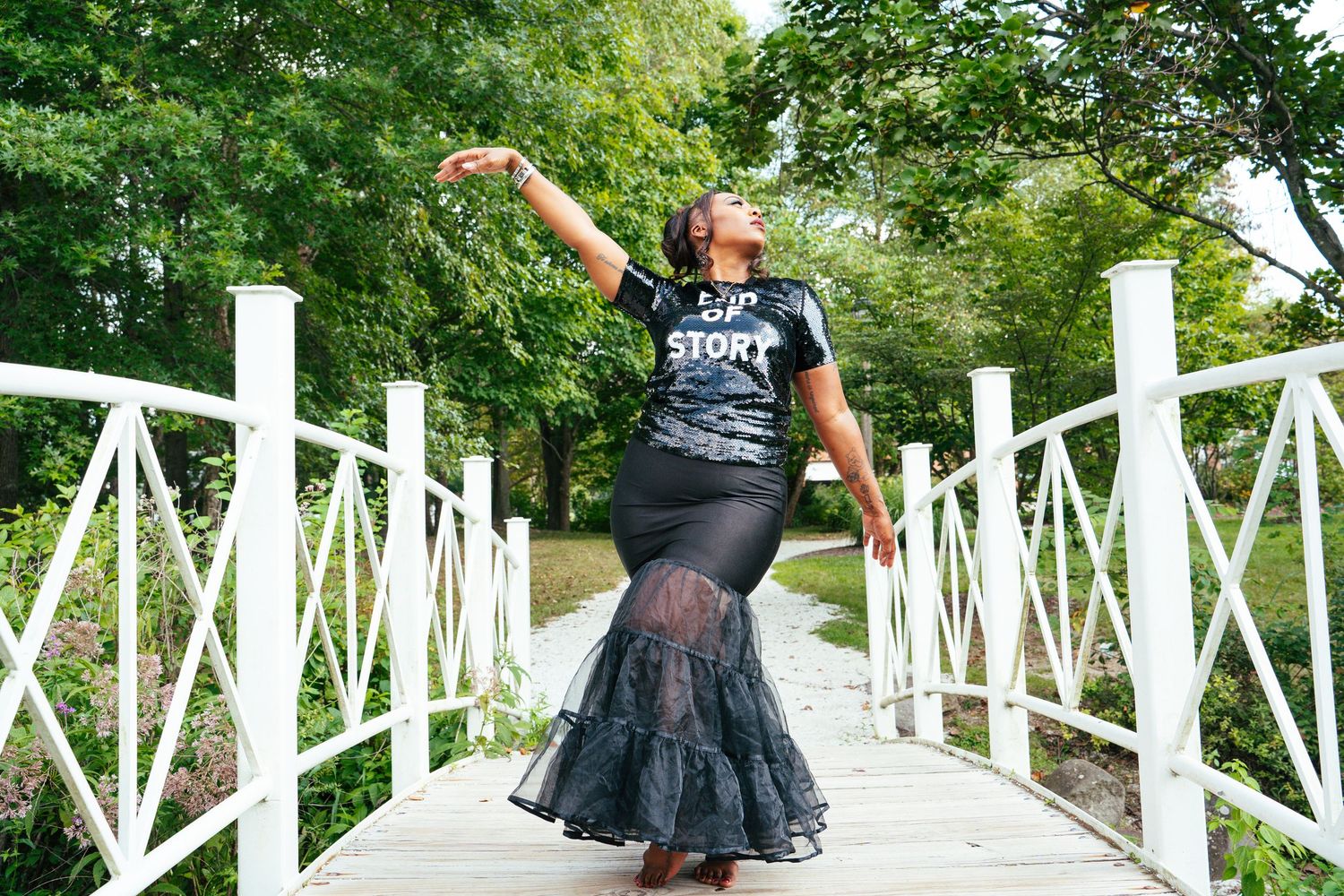 A dancer in a black tutu skirt performs graceful movements on a white bridge surrounded by lush greenery.