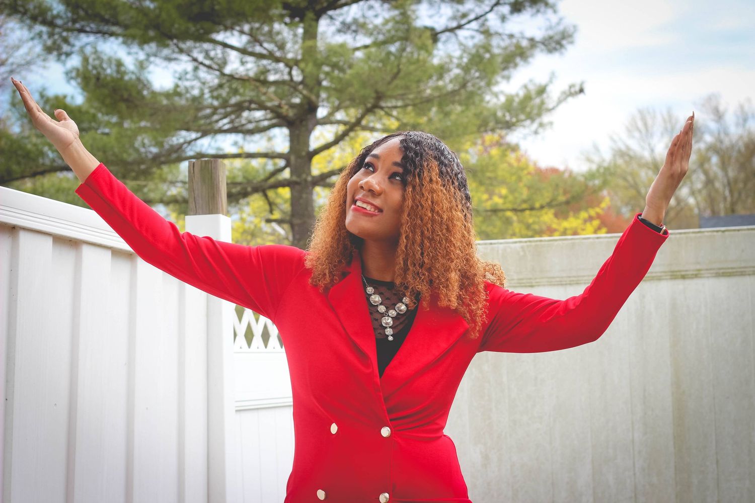 A person in a bright red coat dances and poses joyfully in front of a white fence on a sunny autumn day.