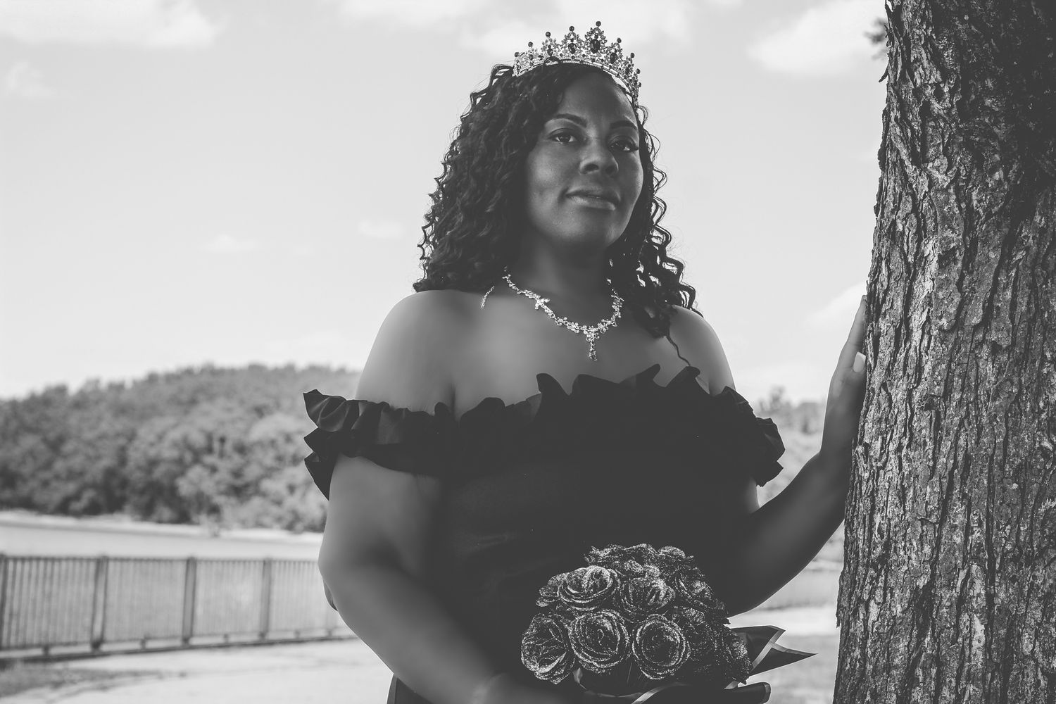 Black and white portrait of a bride in an off-shoulder dress holding a rose bouquet while standing next to a tree.