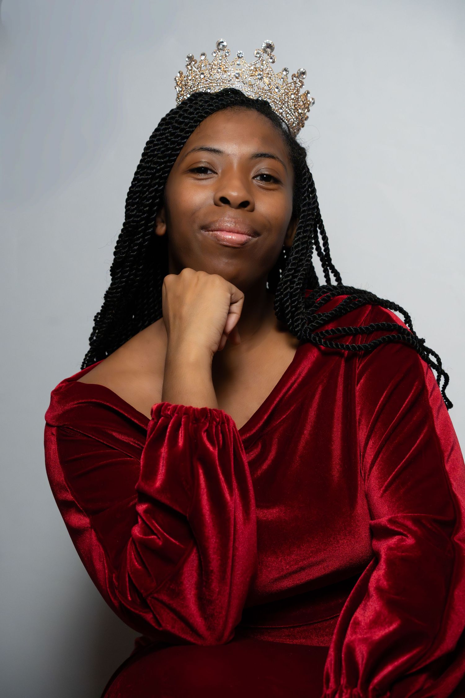 Person in red velvet dress and crystal crown poses elegantly against gray background.