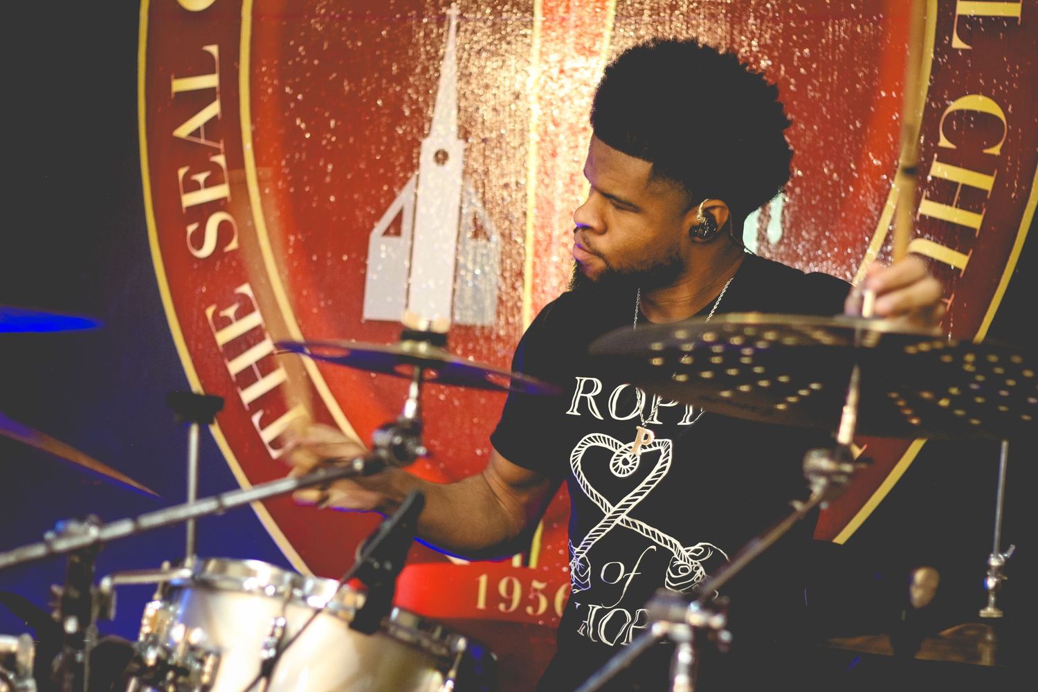 Drummer performs on stage with red lighting and wearing a rock-themed t-shirt.