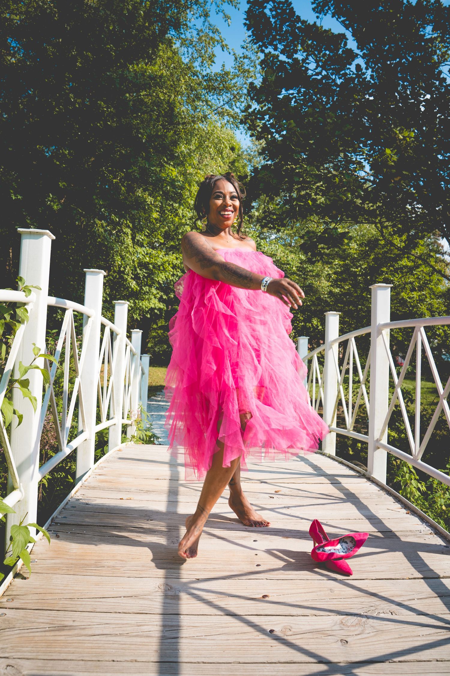 Person in flowing pink tulle dress poses gracefully on a white wooden bridge with red shoes nearby.