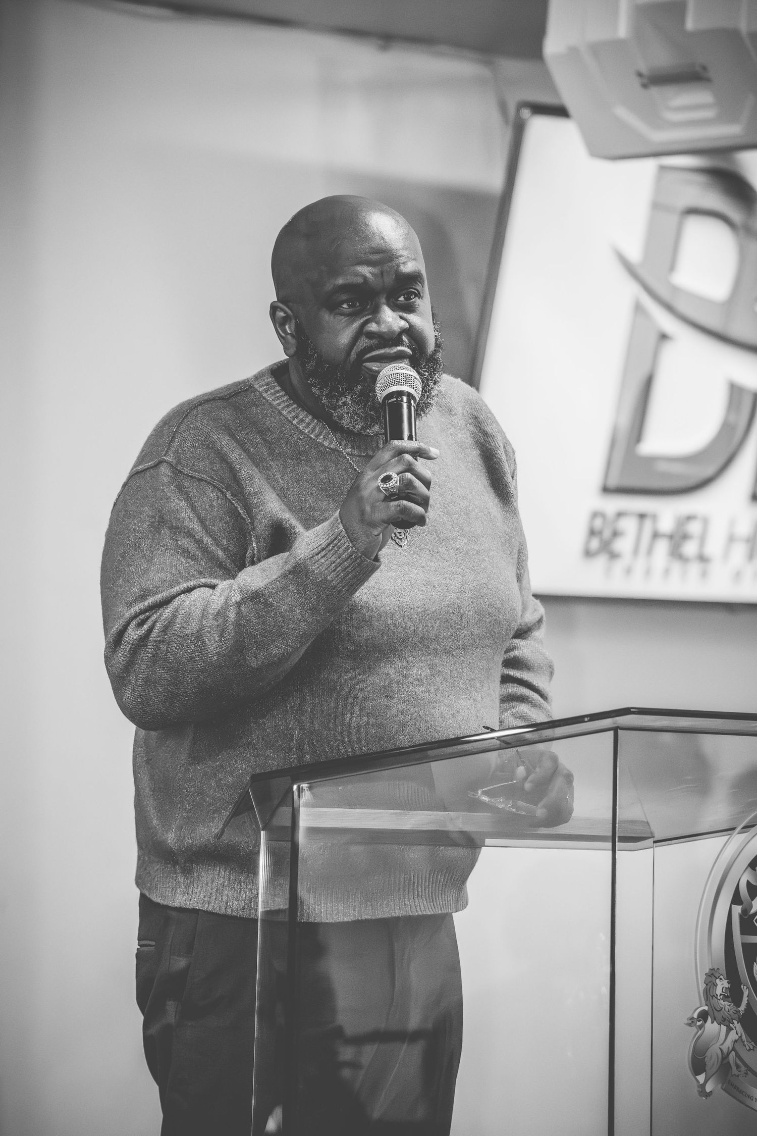 A speaker in a grey sweater addresses an audience from behind a clear podium.