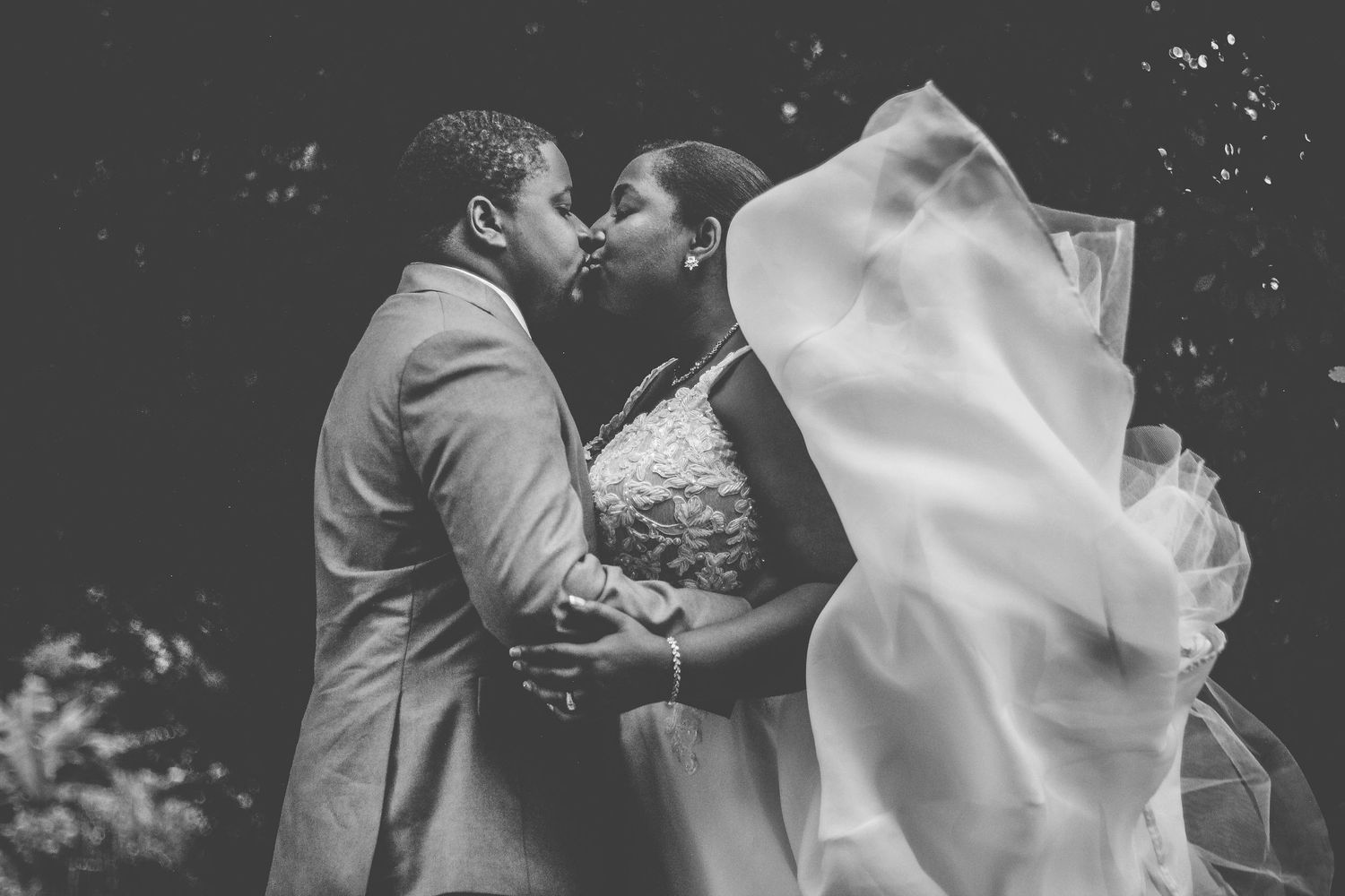 A romantic black and white photo series showing a couple kissing under a flowing veil during their wedding celebration.