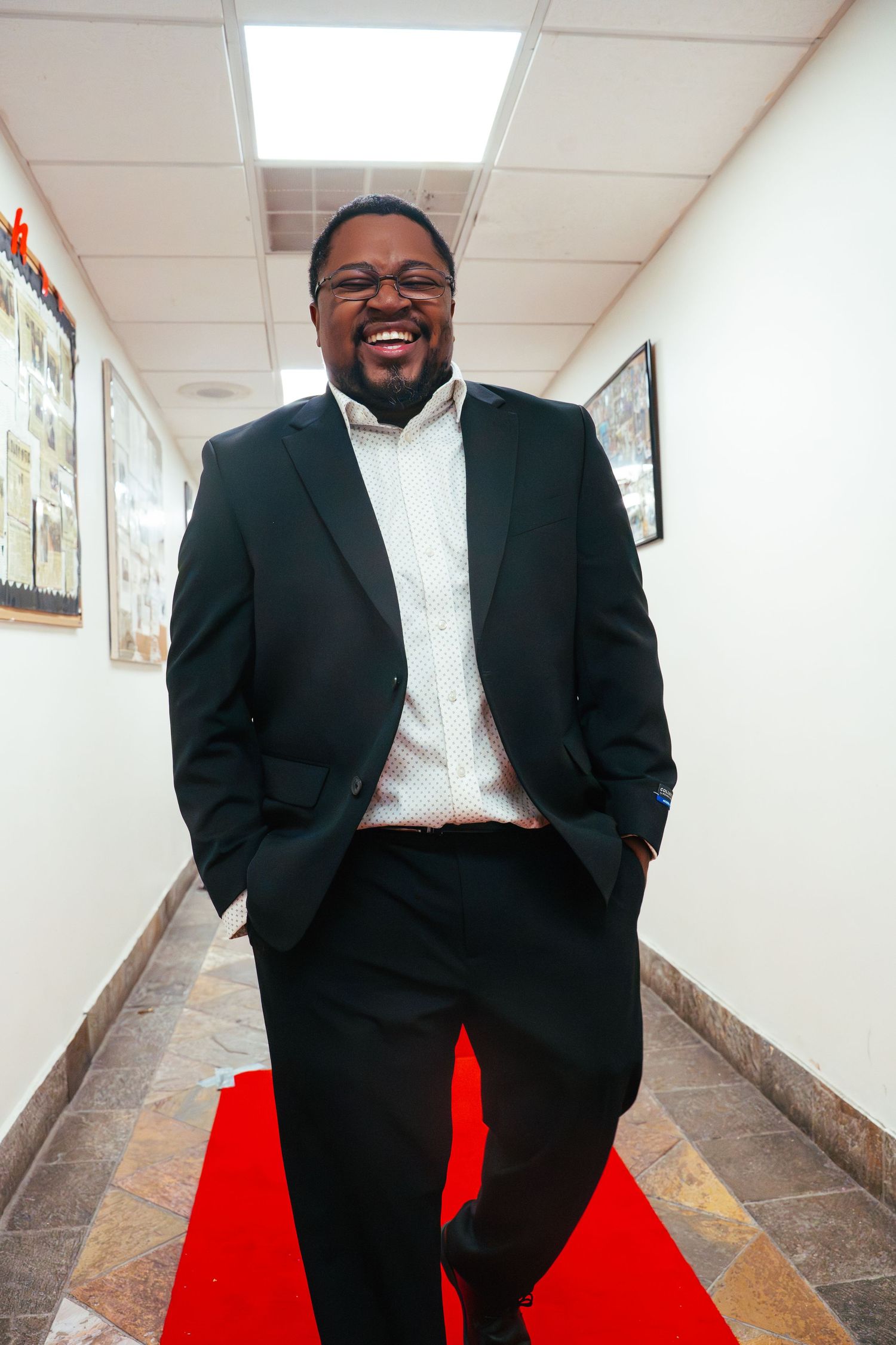 Professional wearing a black suit and white shirt stands smiling in an office hallway with a red carpet.