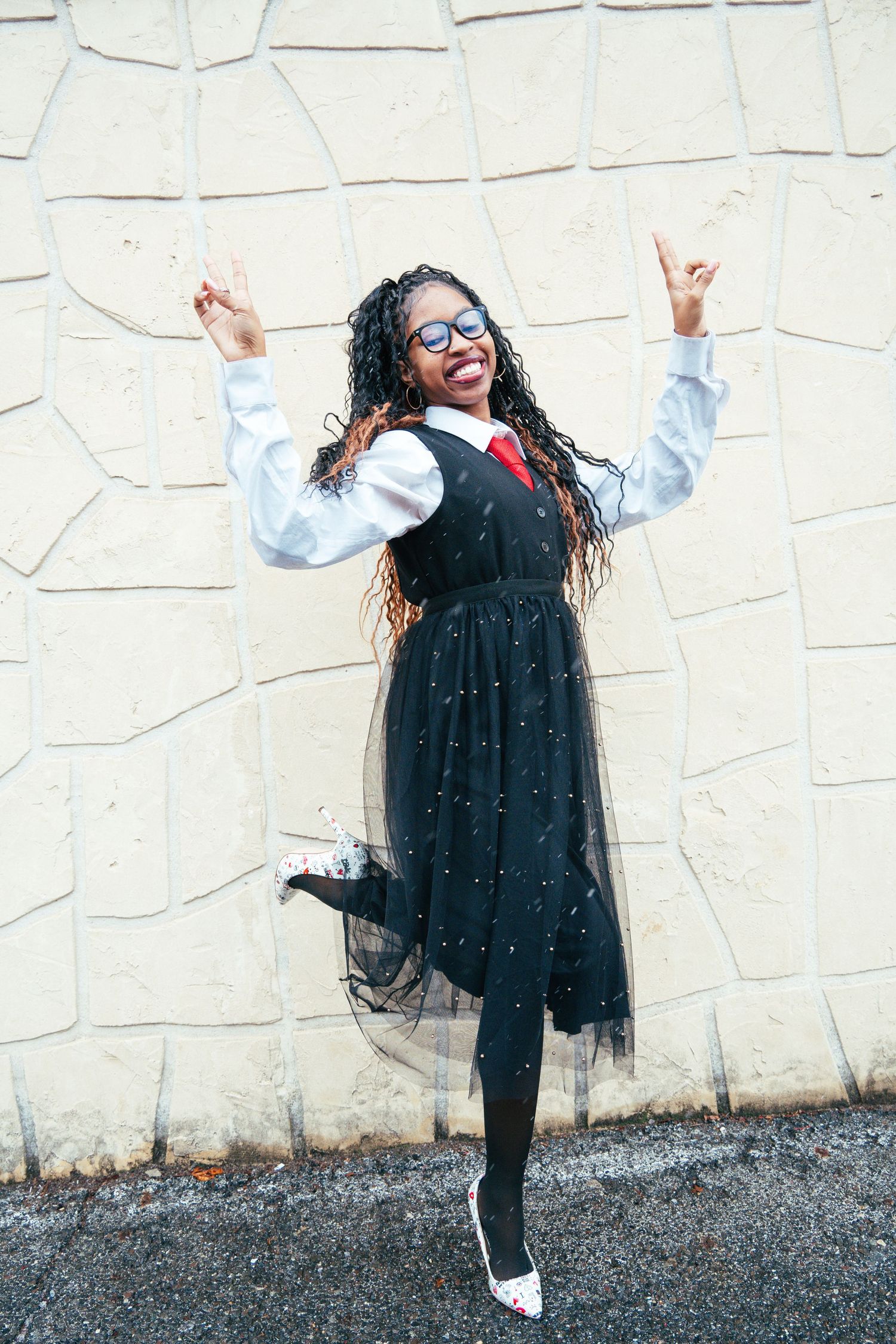 Joyful figure in black dress and white shirt jumps playfully against textured white wall.