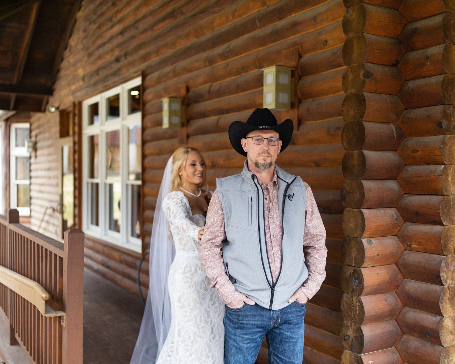 Bride in lace wedding gown and groom in cowboy hat standing outside a rustic log cabin venue.