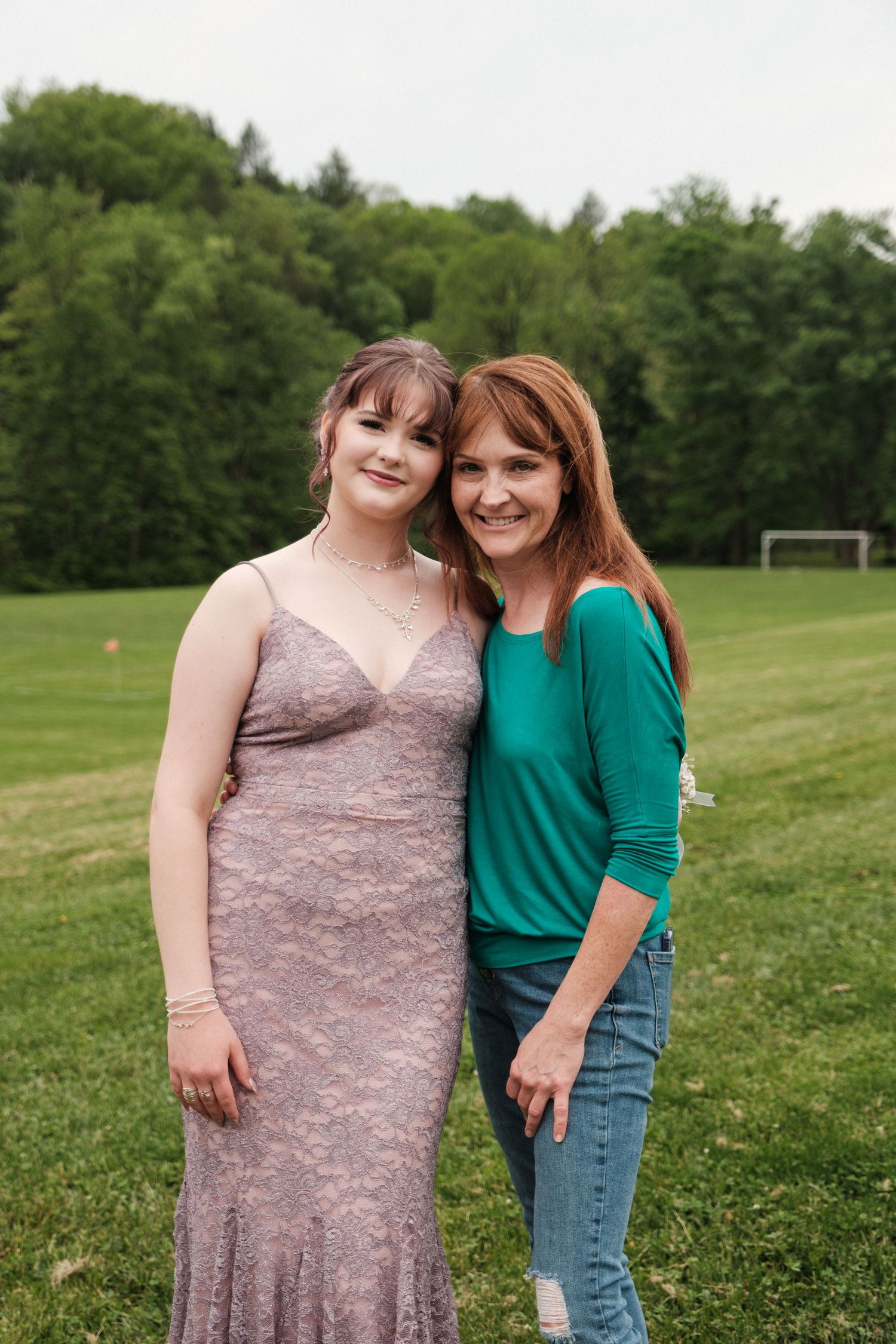 Mother and daughter smile together outdoors on green field before prom celebration.