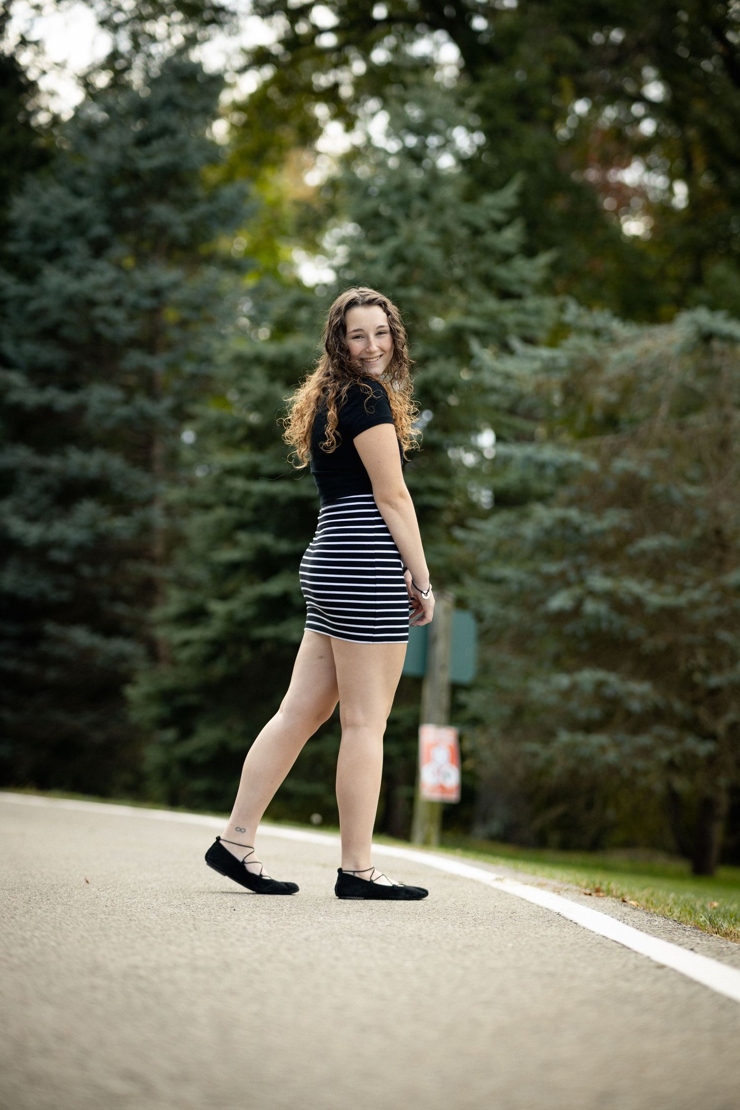 Young woman in striped mini skirt and black top posing on a tree-lined road in autumn.