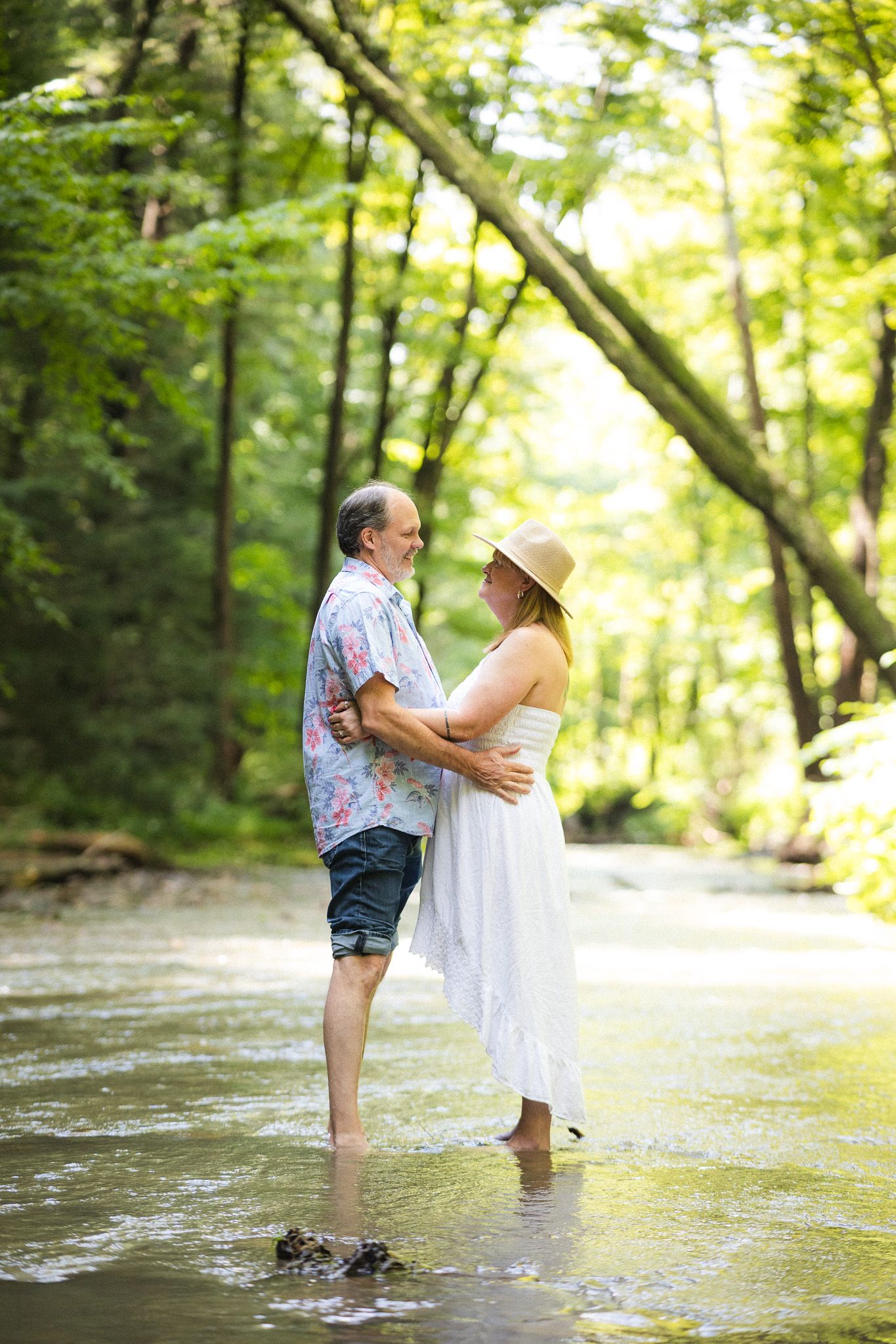 Happy mature couple embracing barefoot in a shallow forest creek surrounded by lush green trees.