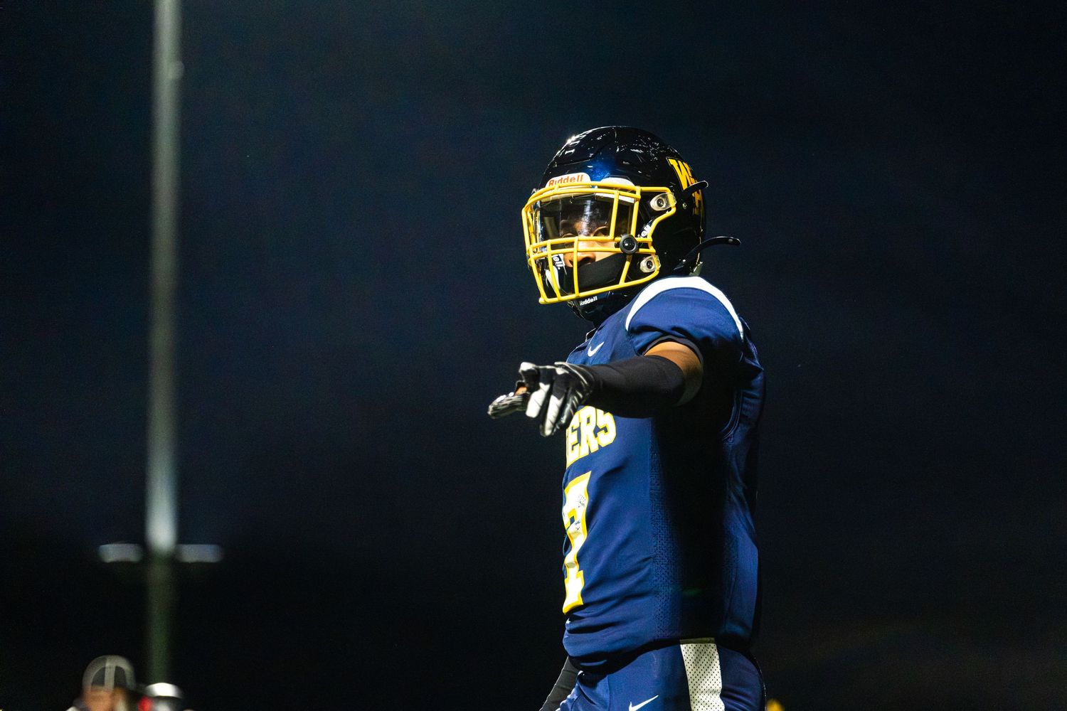 A football player in blue and gold uniform points on a dark night game field.