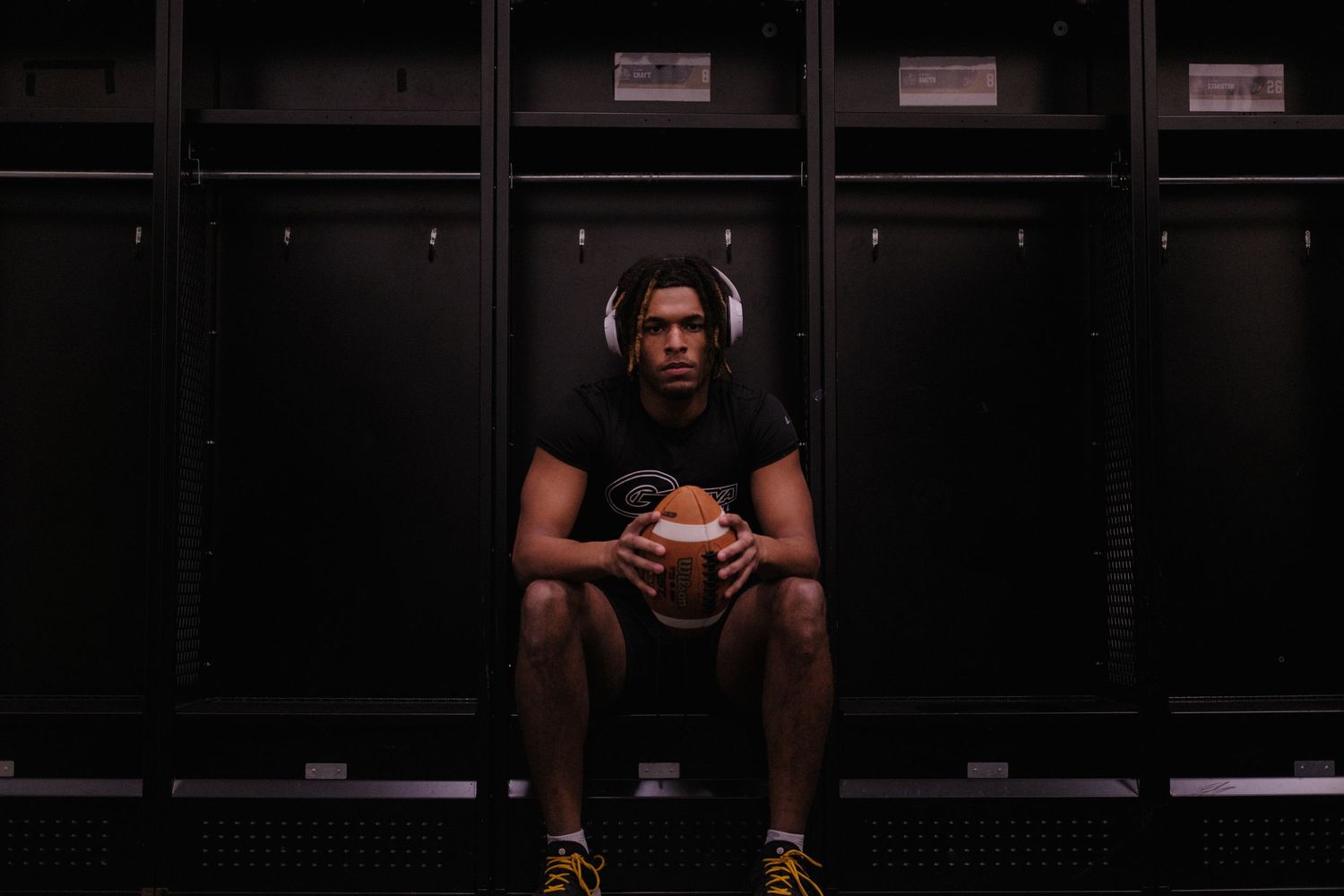Male athlete sitting in dark locker room holding a football wearing headphones.
