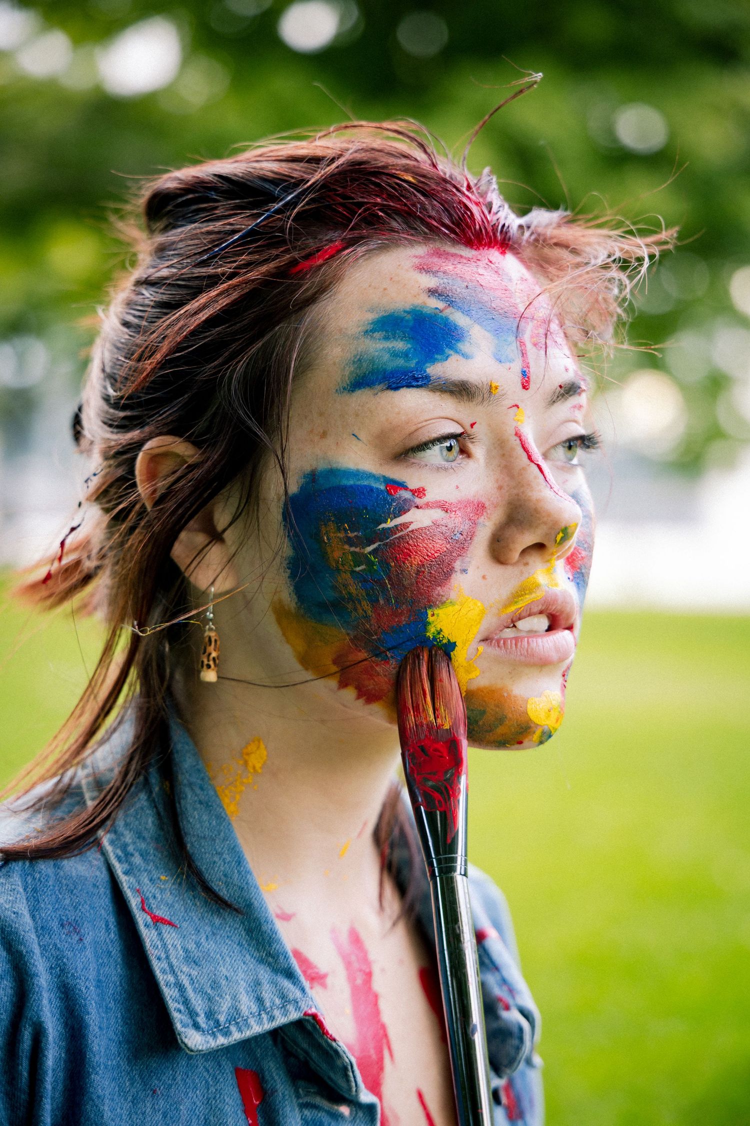 Artistic woman with multicolored paint on face holds paintbrush outdoors in park.