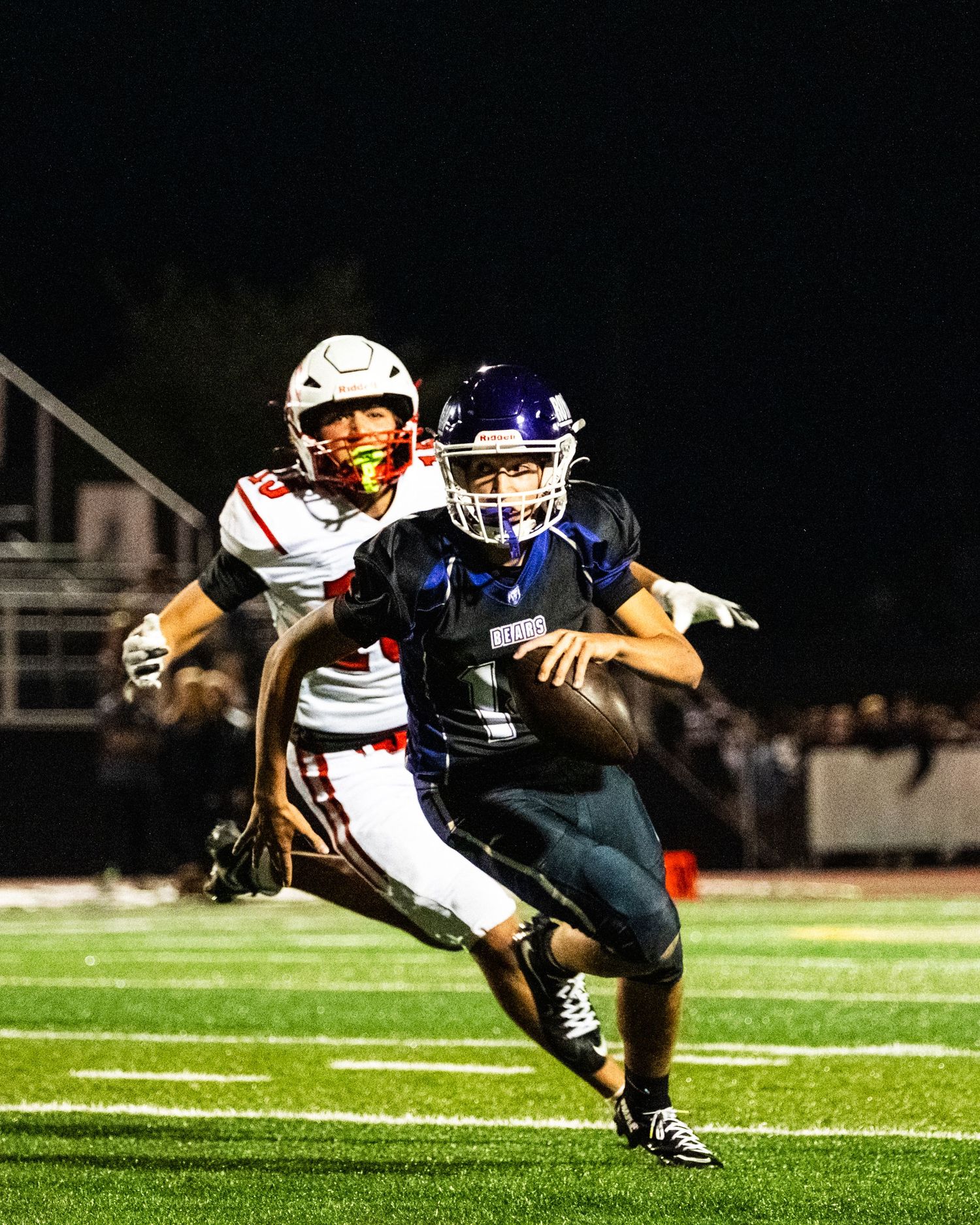 High school football player in blue uniform scrambles away from red-jersey defender during night game.