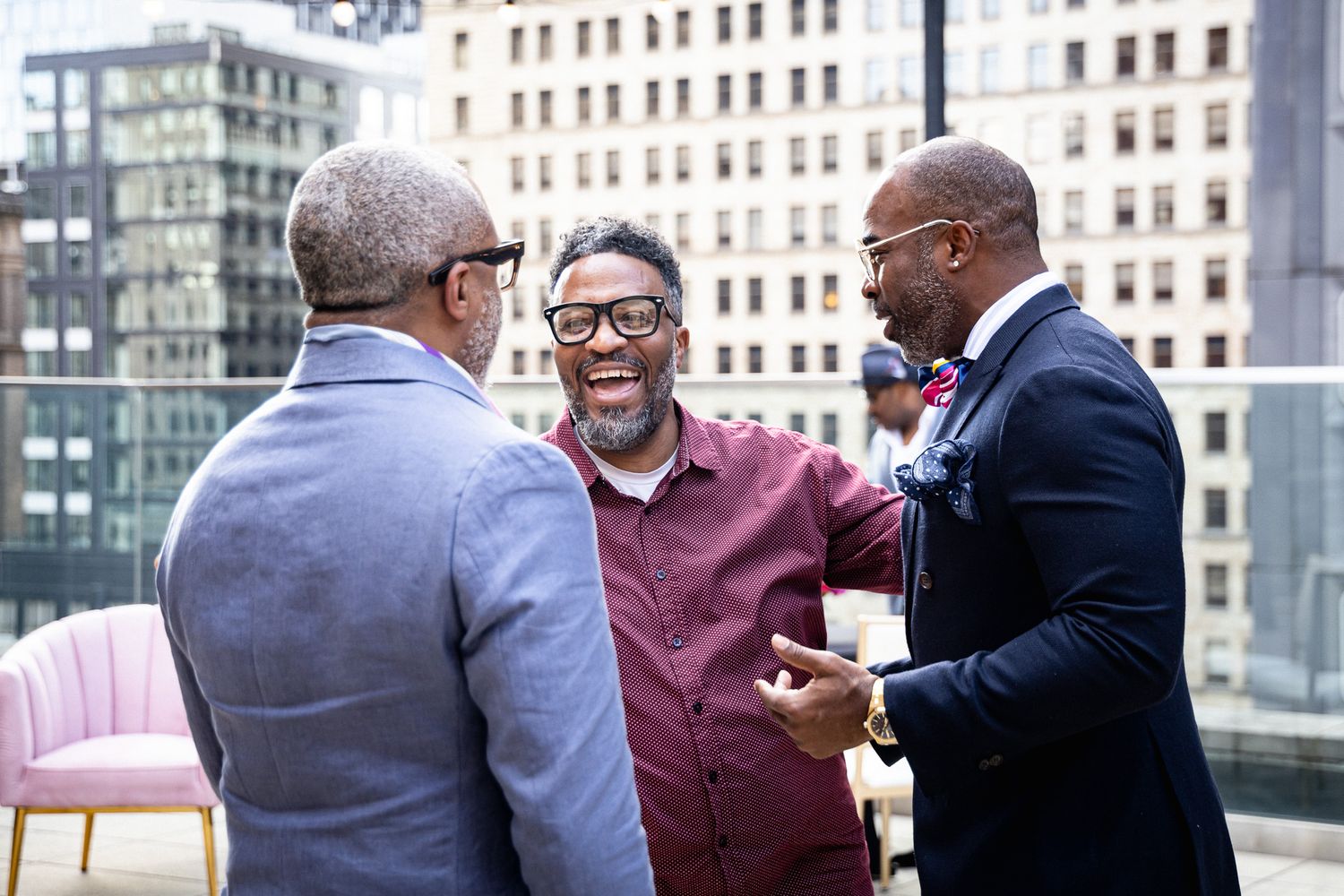 Three men laughing and embracing on urban rooftop terrace with city skyline in background.